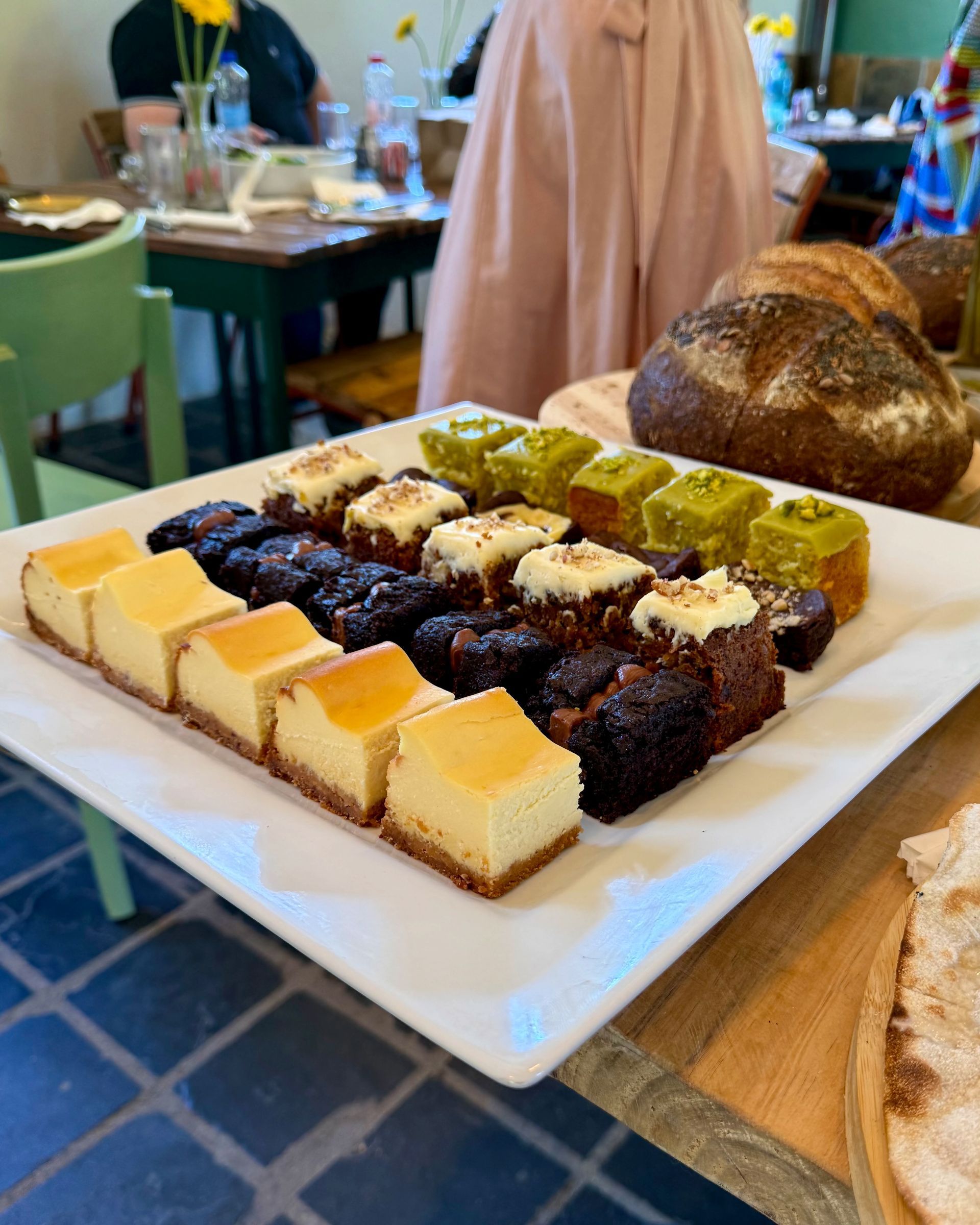 A tray of baked goods: cheesecake, brownies, and pistachio squares, on a table. People and bread are in the background.