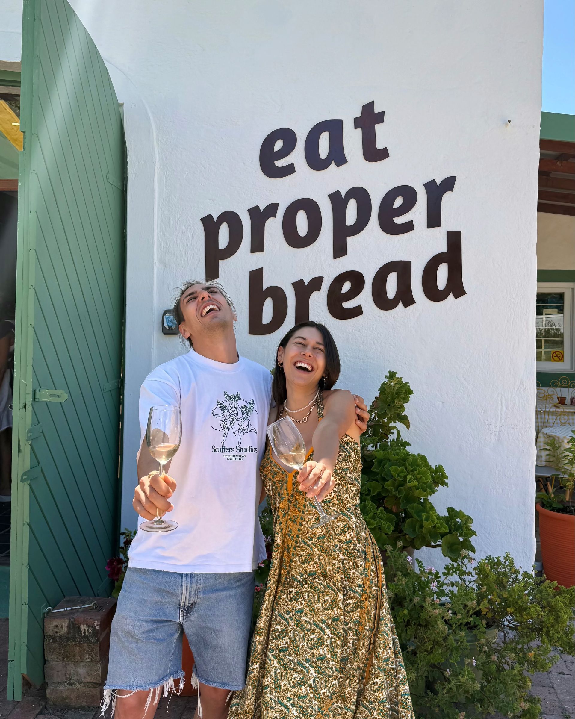 Two people smiling, toasting drinks, in front of a sign that reads 