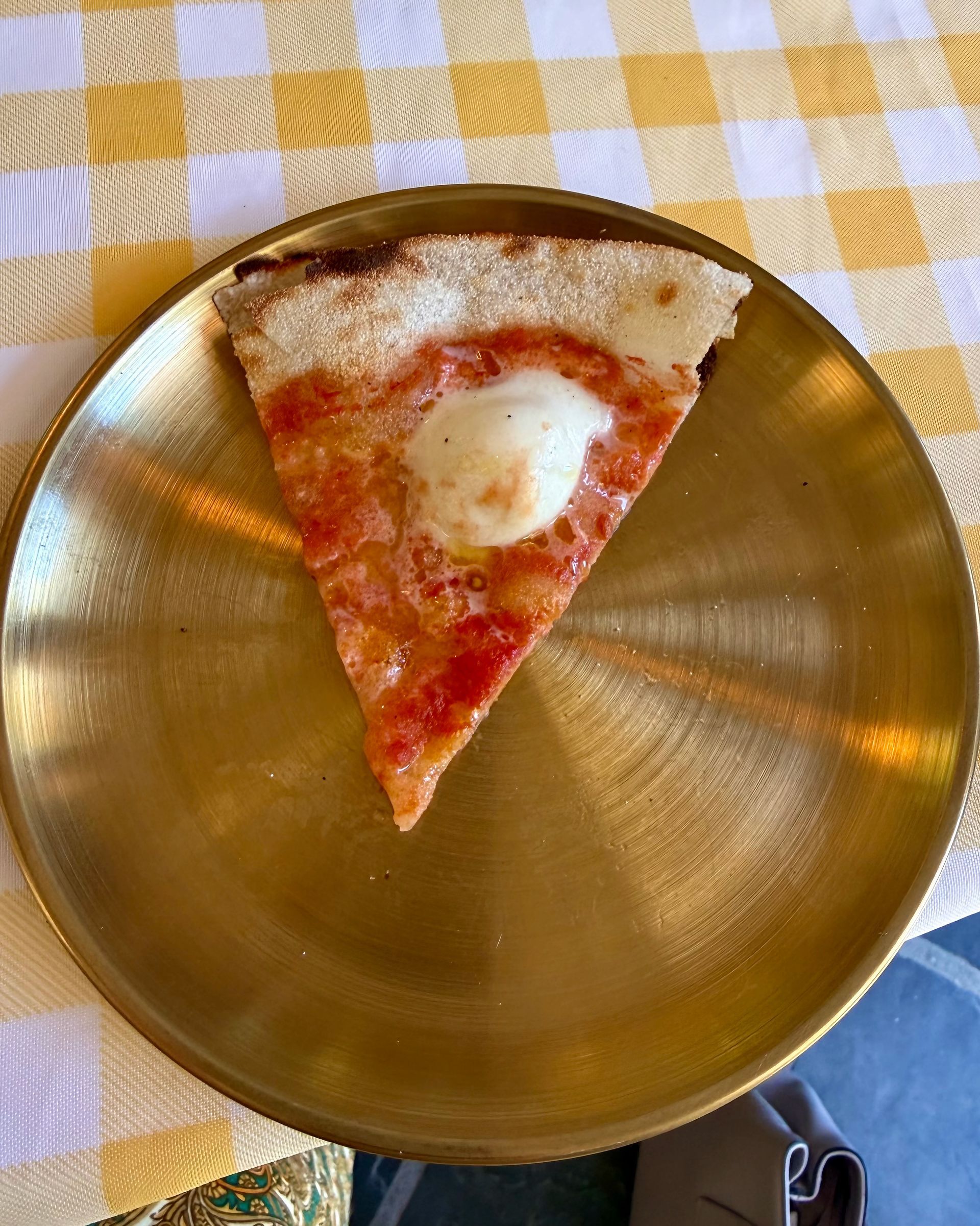 Slice of pizza on a gold plate, set on a yellow checkered tablecloth.