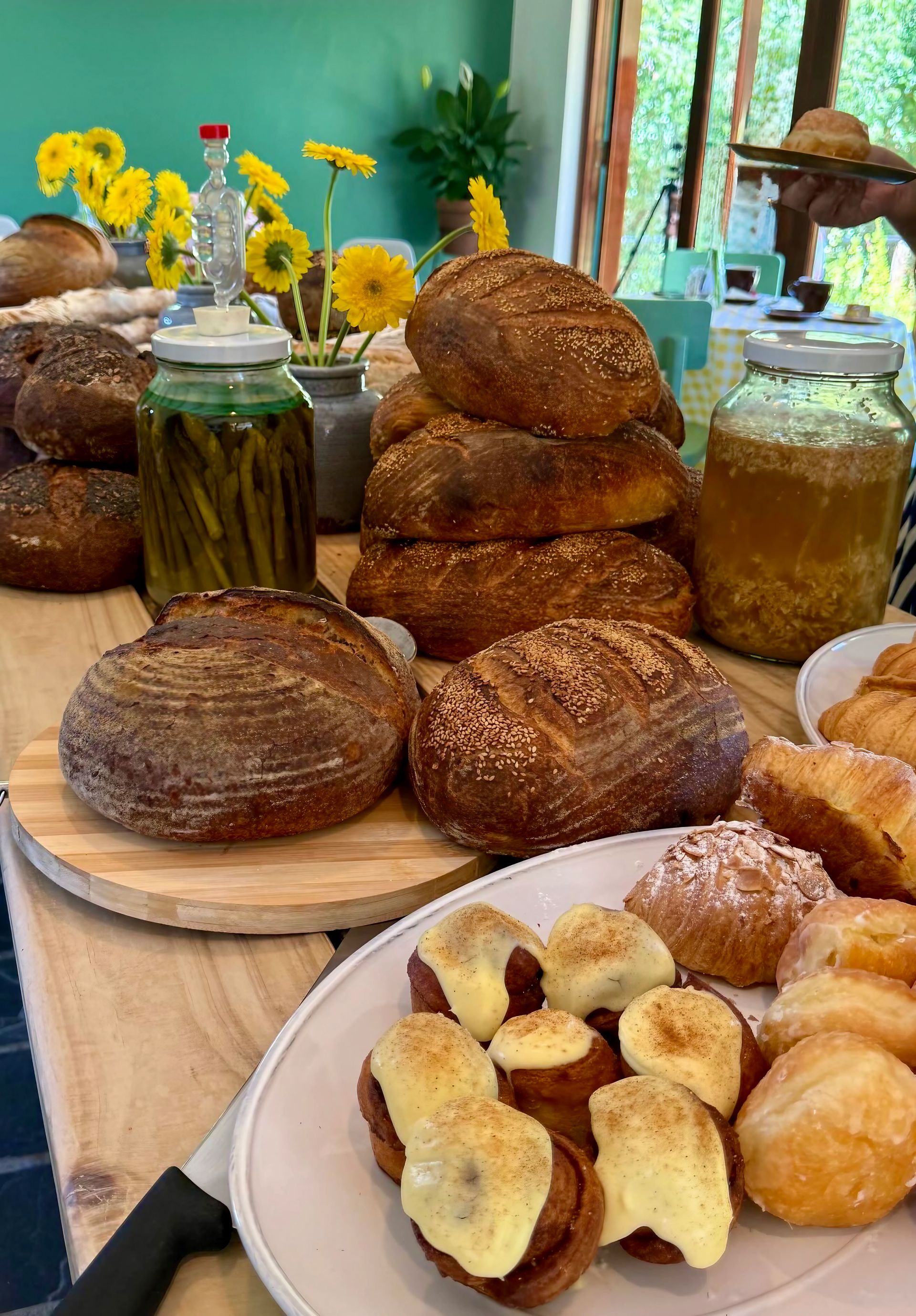 Table of breads, jars, and pastries, with yellow flowers in background.