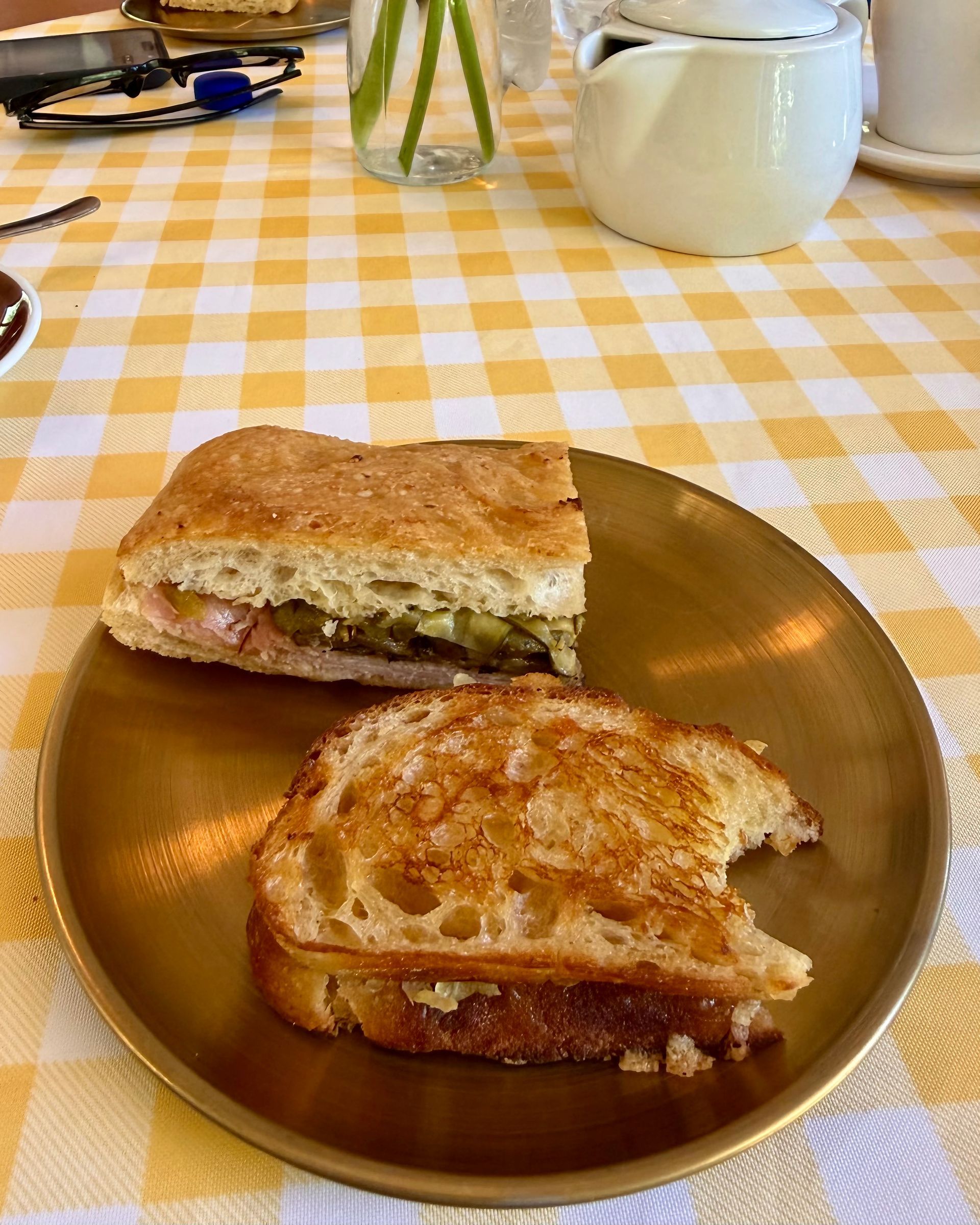 Two sandwiches on a gold plate on a yellow checkered tablecloth. One sandwich is cut in half, the other has a bite taken out.