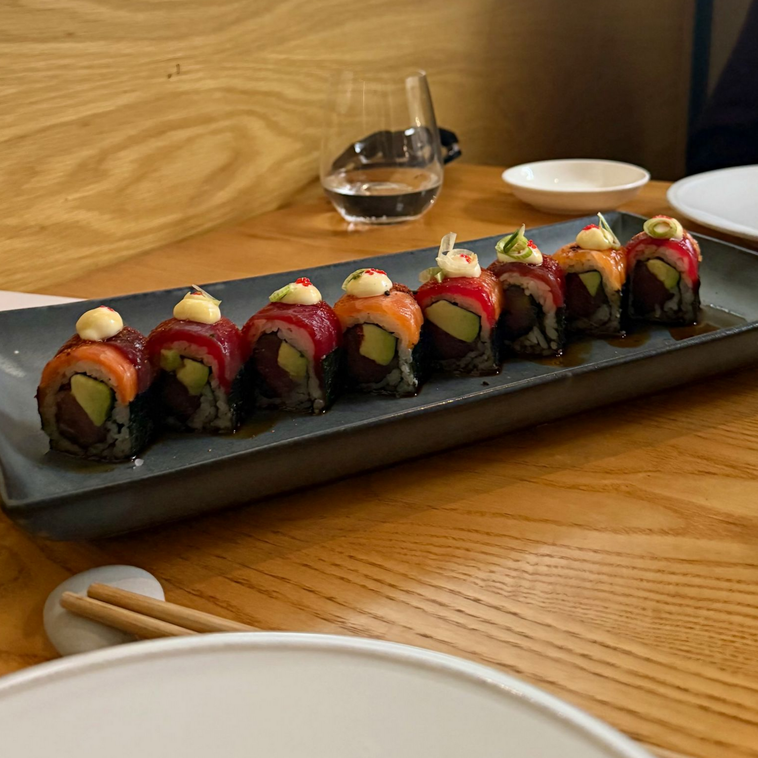 Sushi rolls on a rectangular plate on a wooden table. Toppings include seafood and sauce.