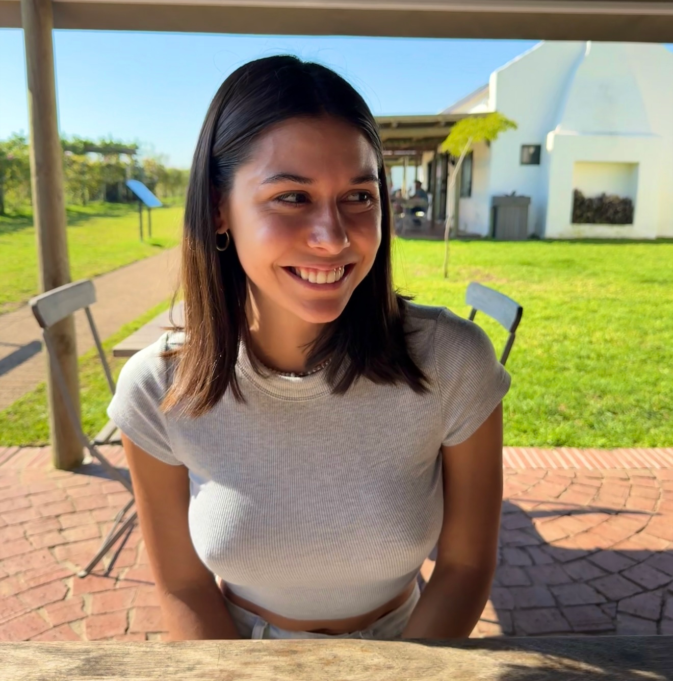Woman with a big smile outdoors at a cafe, sitting in front of a white building, sunny day.