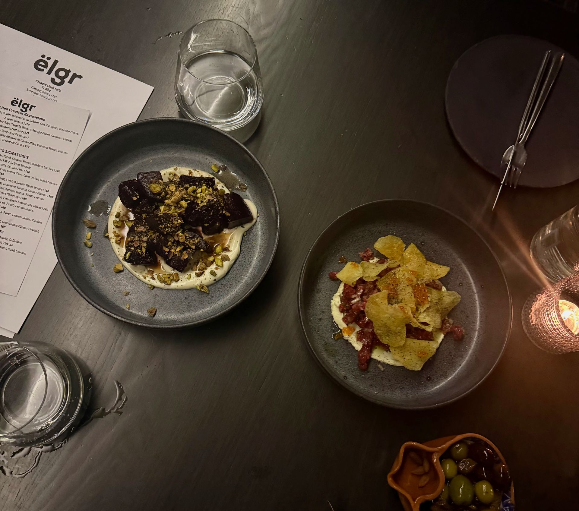 Two dishes on plates with a menu, drinks, and a candle on a dark table.