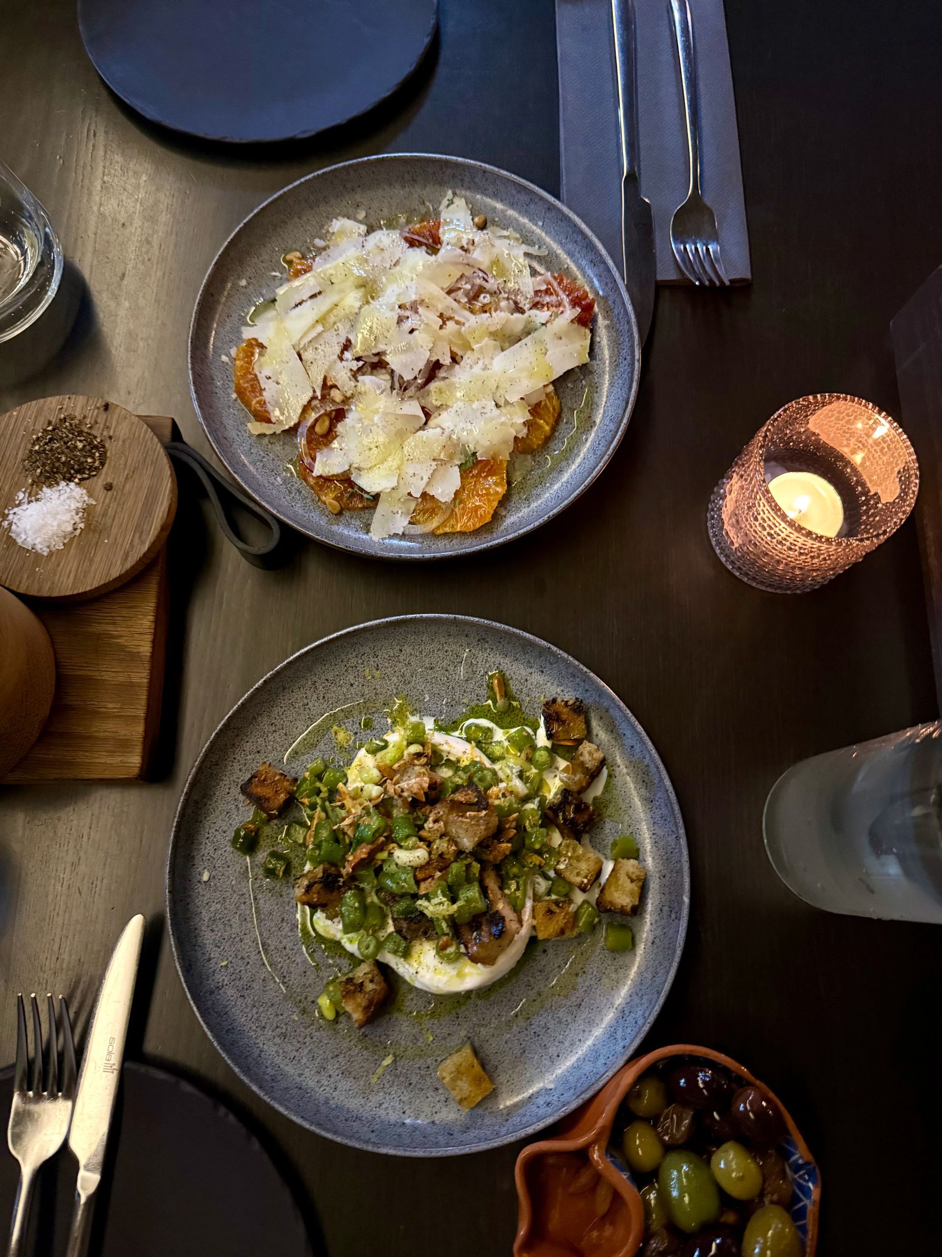 Overhead view of two plates with food, candle, and olive bowl on a dark table.