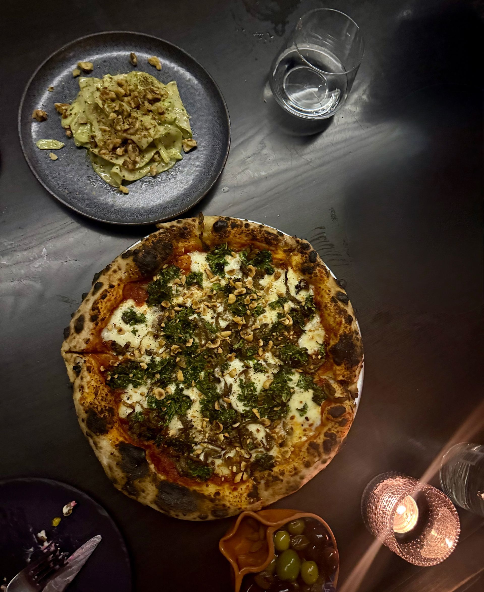 Overhead shot of a pizza, pasta, glass of water, and olives on a dark table.