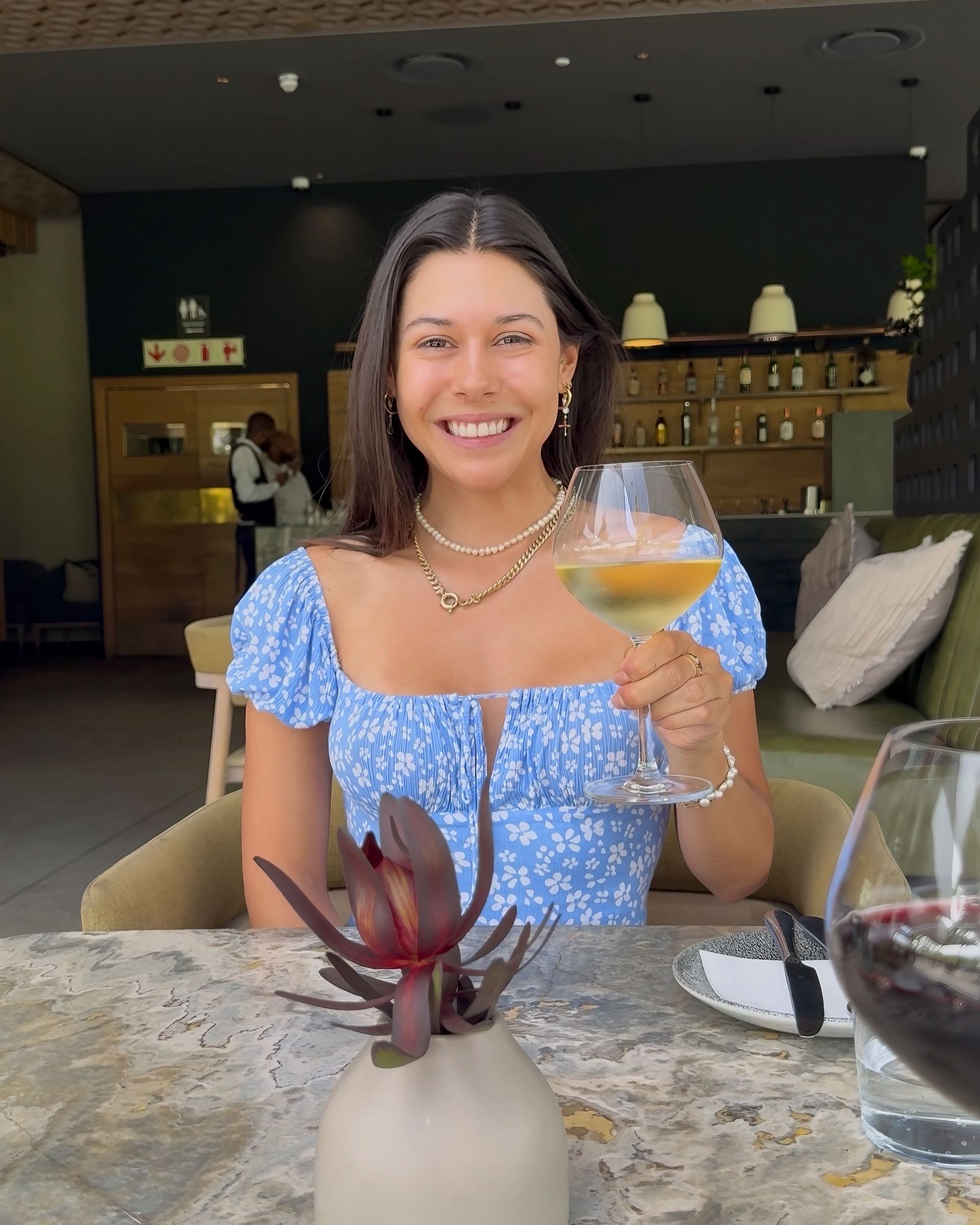 Woman smiling, holding wine glass at a restaurant, floral blue top, seated at a table.