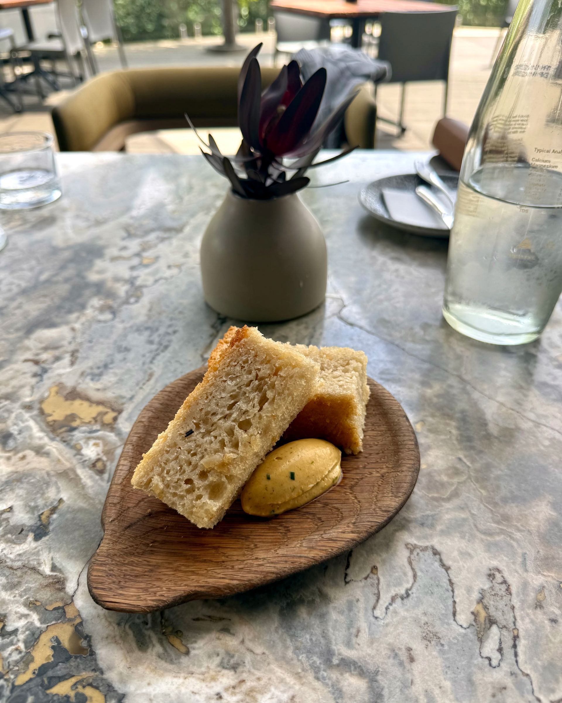 Bread slices with butter on a wooden tray, alongside a water glass and vase with purple flower on a marble table.