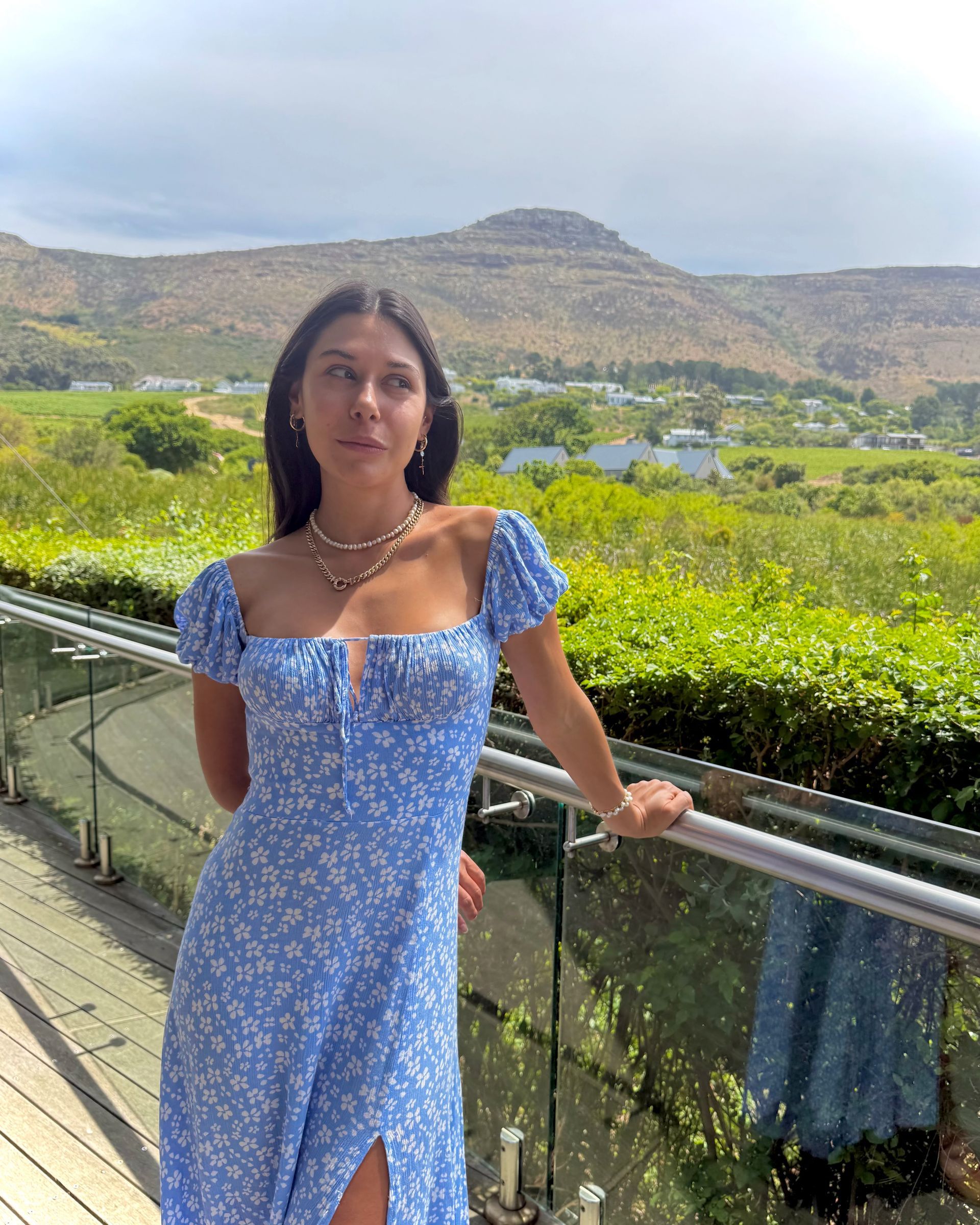 Woman in a blue floral dress poses on a balcony overlooking a scenic landscape.