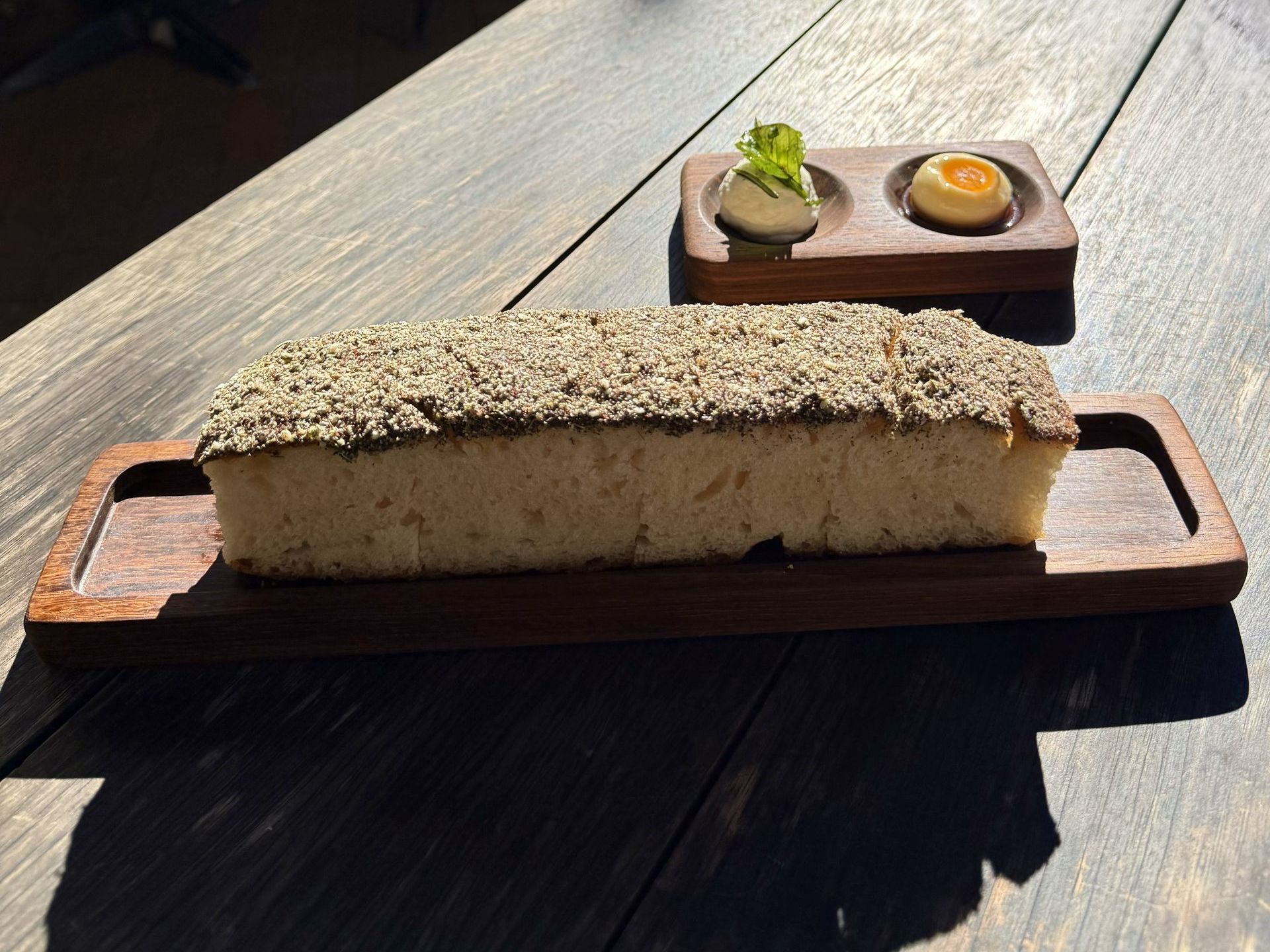 A wooden tray with seeded bread and a side of butter and a round spread.