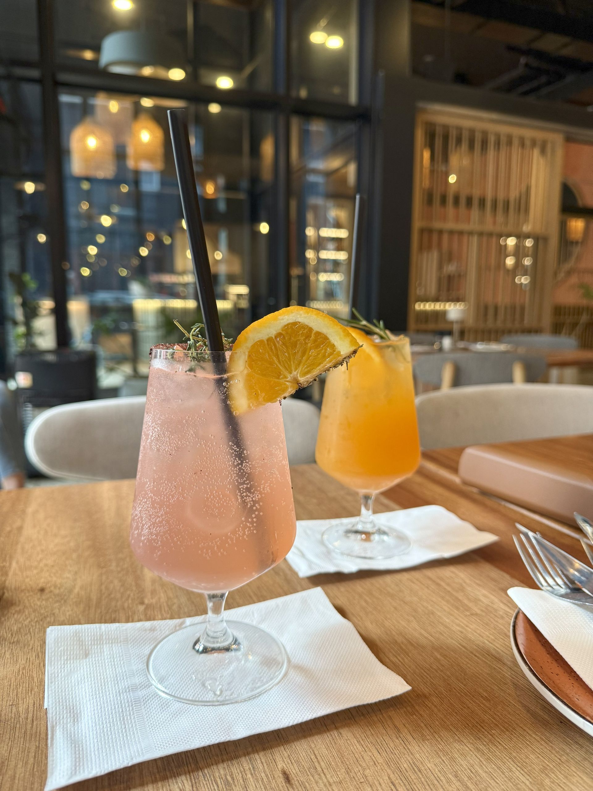 Two cocktails, one pink and one orange, garnished with fruit, on a table in a restaurant.