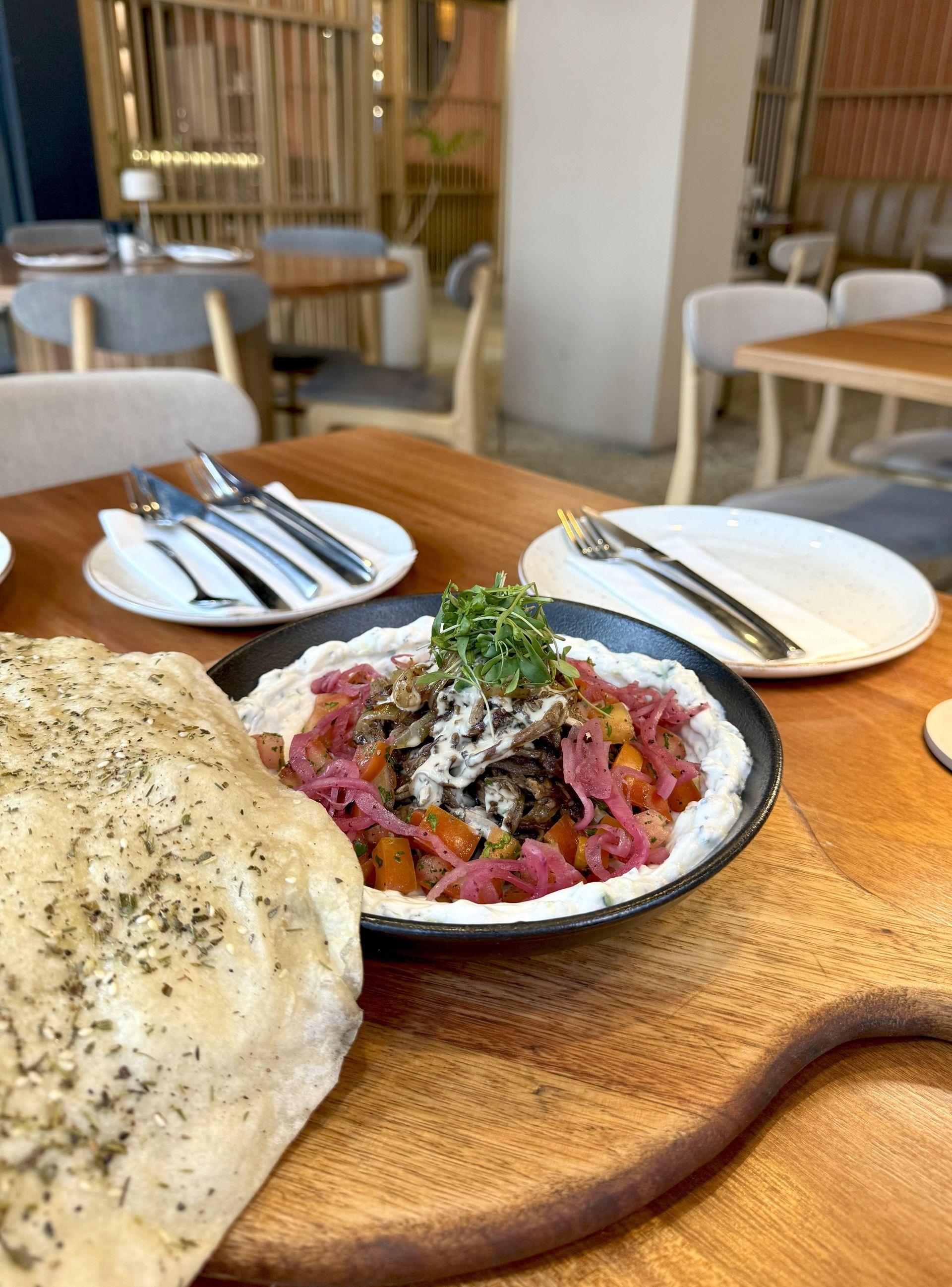 Hummus dish topped with meat and veggies, served with pita bread, on a wooden table. Restaurant interior.
