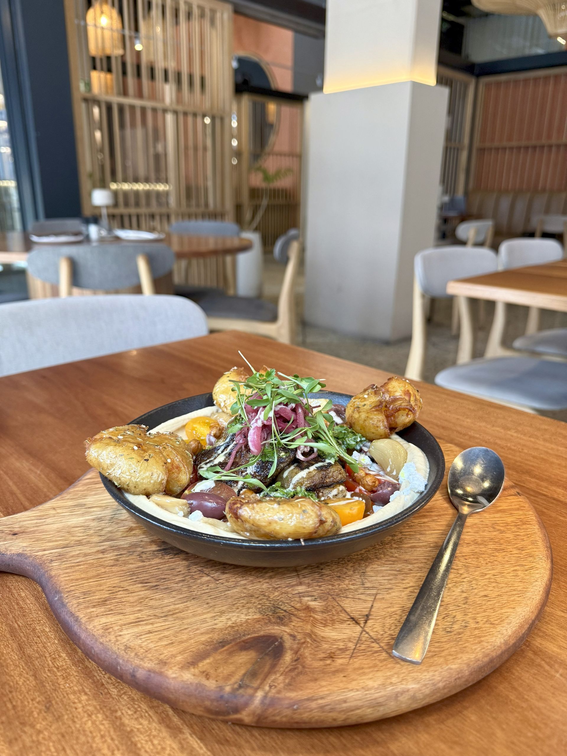 Hummus bowl with potatoes, vegetables, and greens on a wooden board in a restaurant setting.