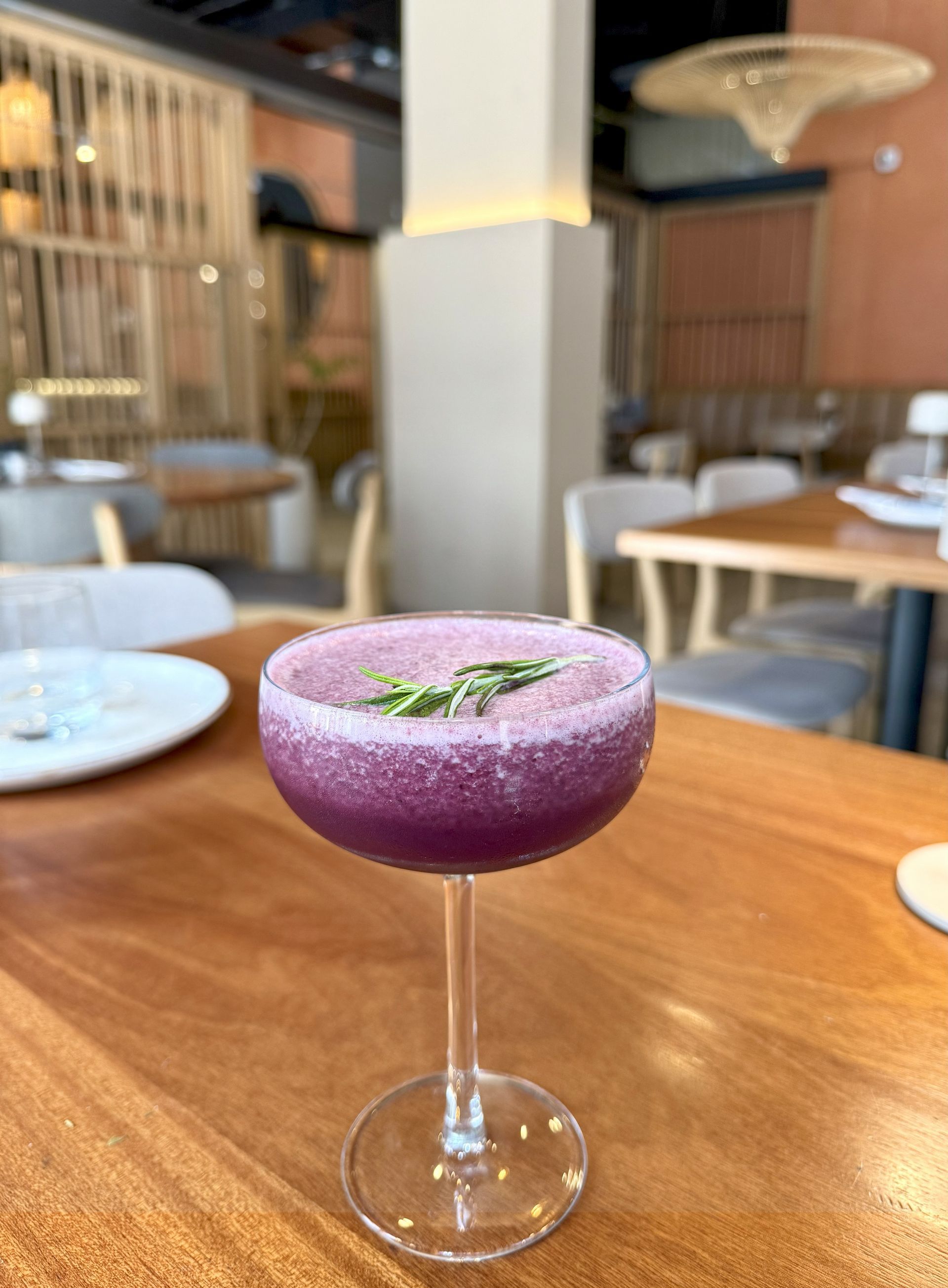 Purple cocktail in a stemmed glass, rosemary garnish, on a wooden table in a restaurant.