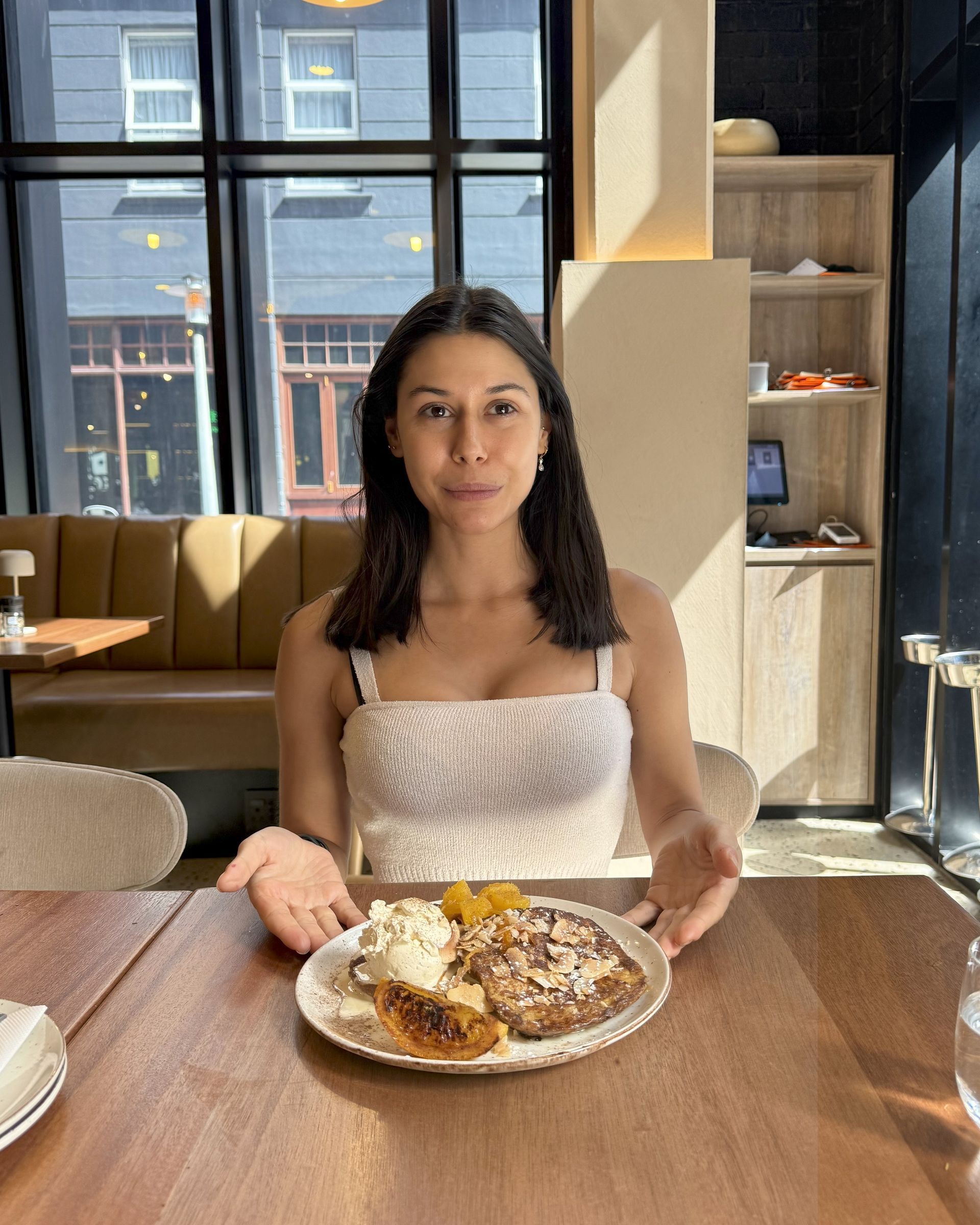 Woman smiling, sitting at a table with a dessert plate in front of her. Sunlight, restaurant setting.