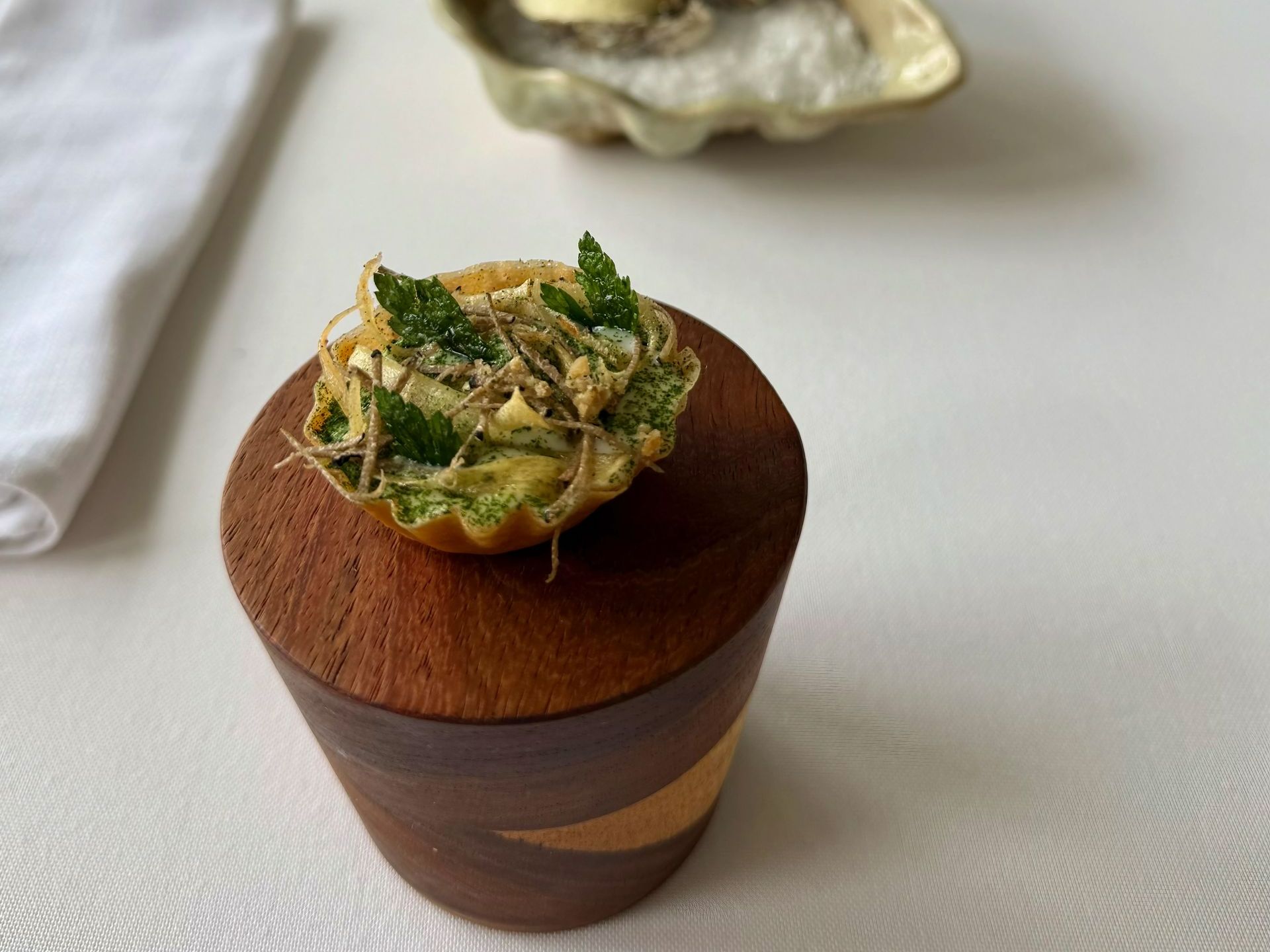 A small, golden pastry with herbs sits on a wooden stand, next to an oyster on a shell.