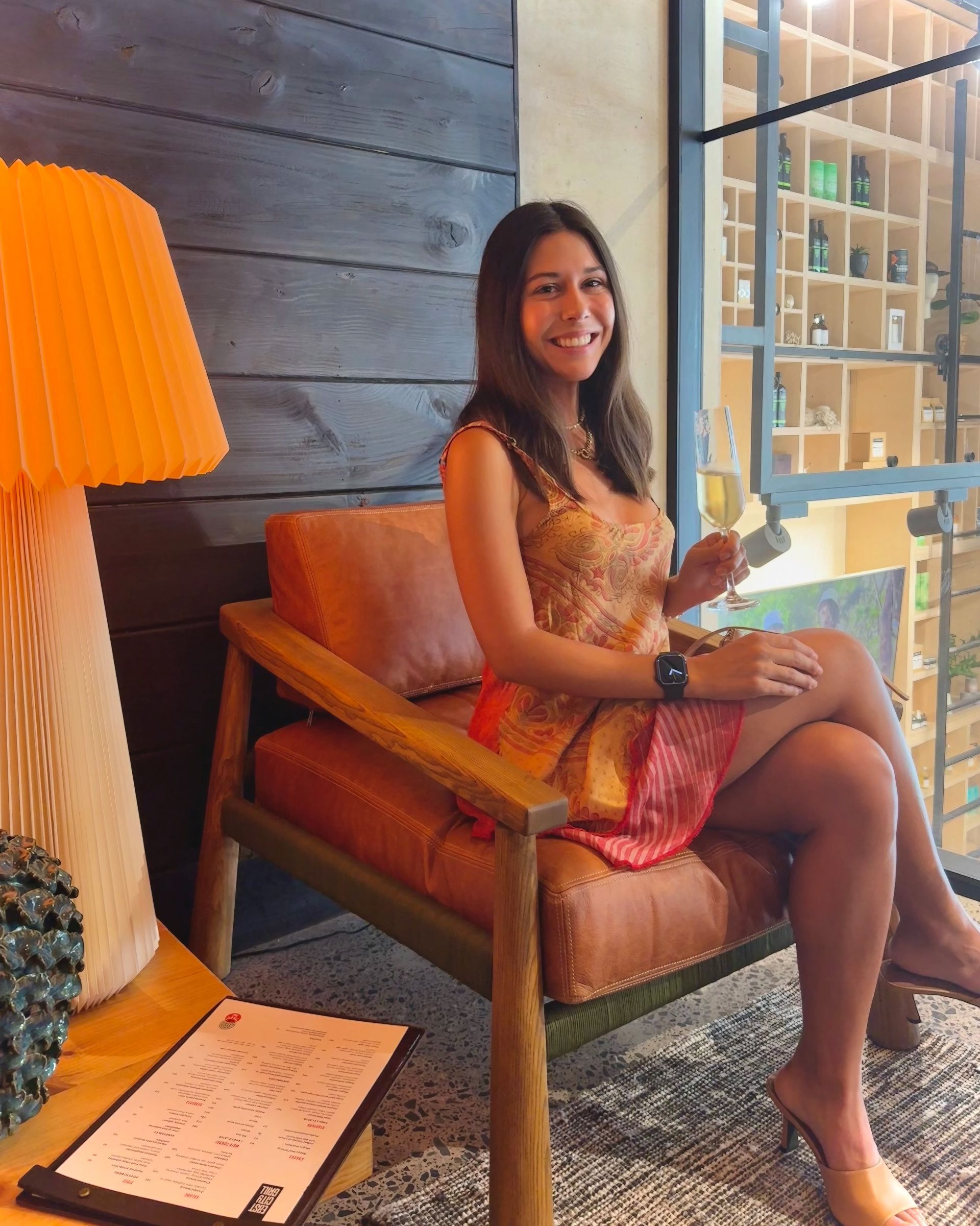 Woman in a dress, holding a glass, seated in a chair, smiling. Interior setting with a lamp and shelves.