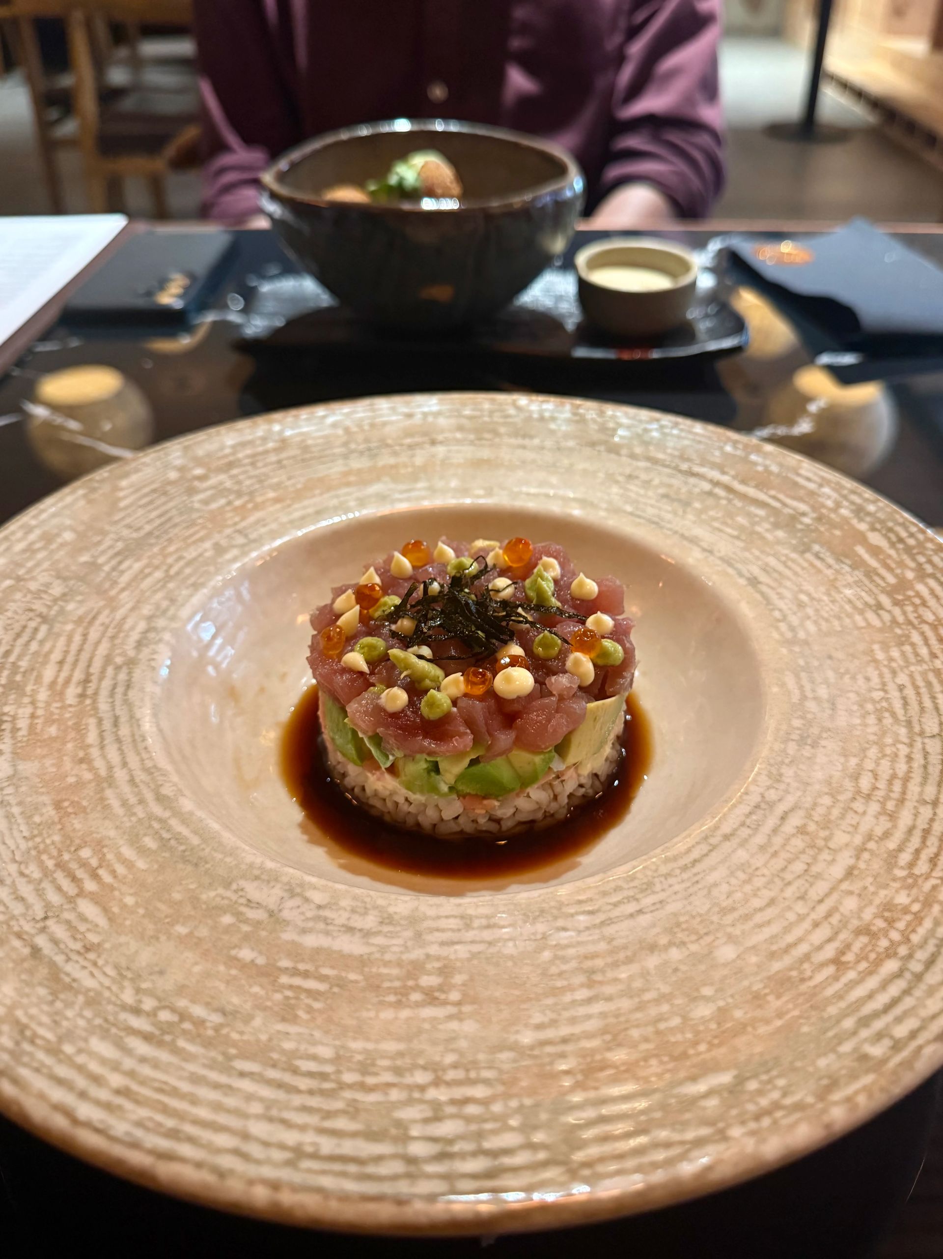 Sushi tower on a plate with sauce, person in background, and bowl of food.