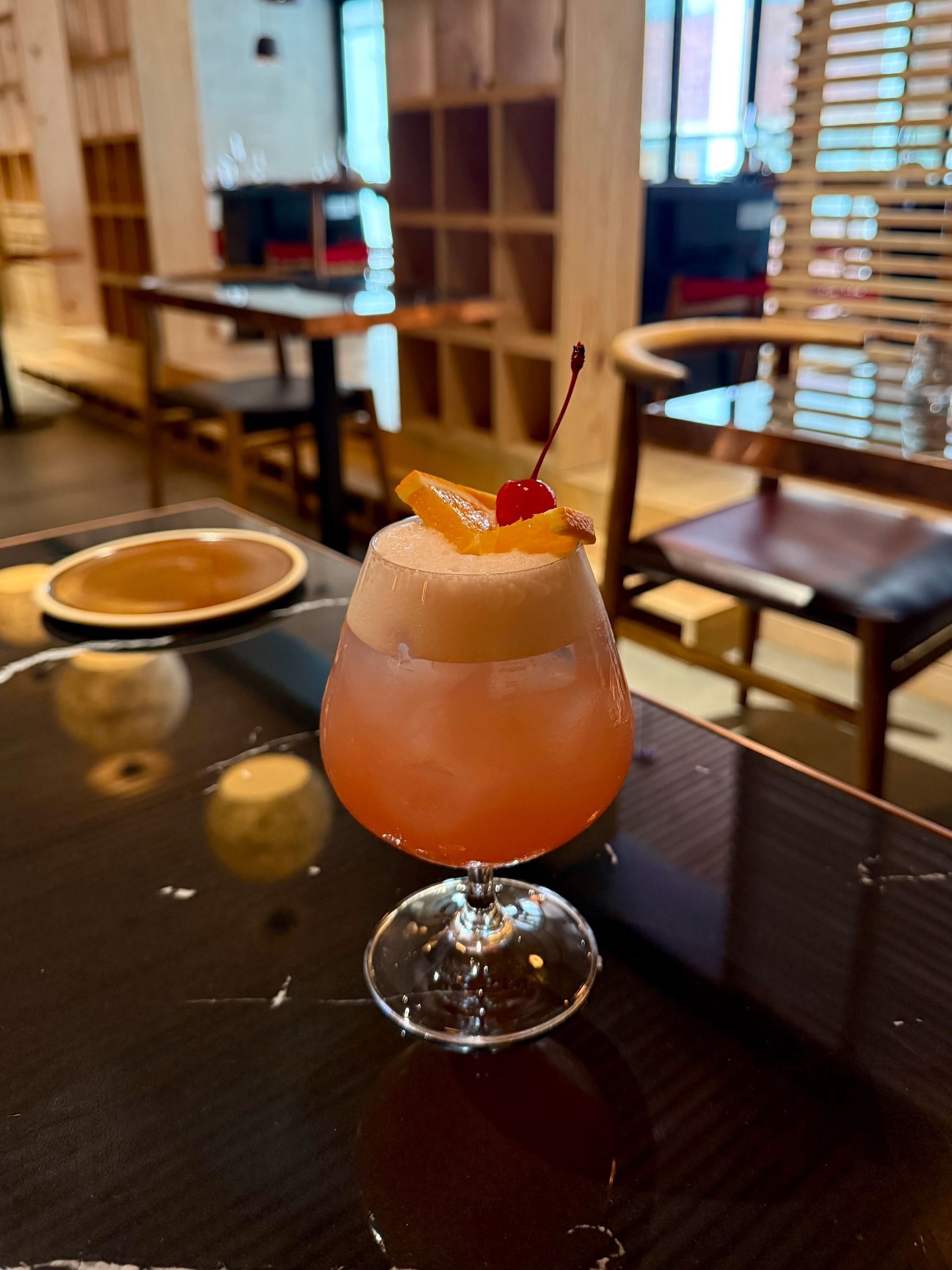 Cocktail in a stem glass with foam top, garnished with an orange slice and cherry, on a black table. Restaurant interior in background.