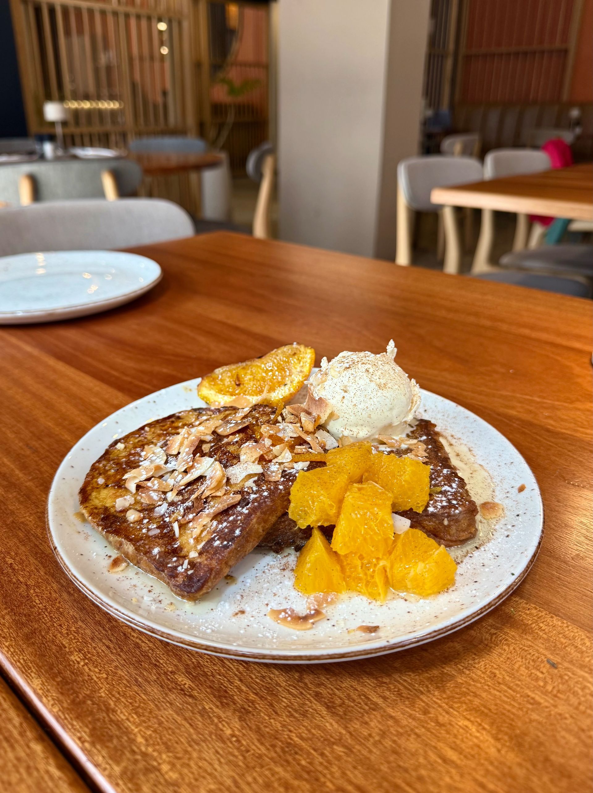French toast with orange slices, cream, and almonds on a white plate, set on a wooden table.