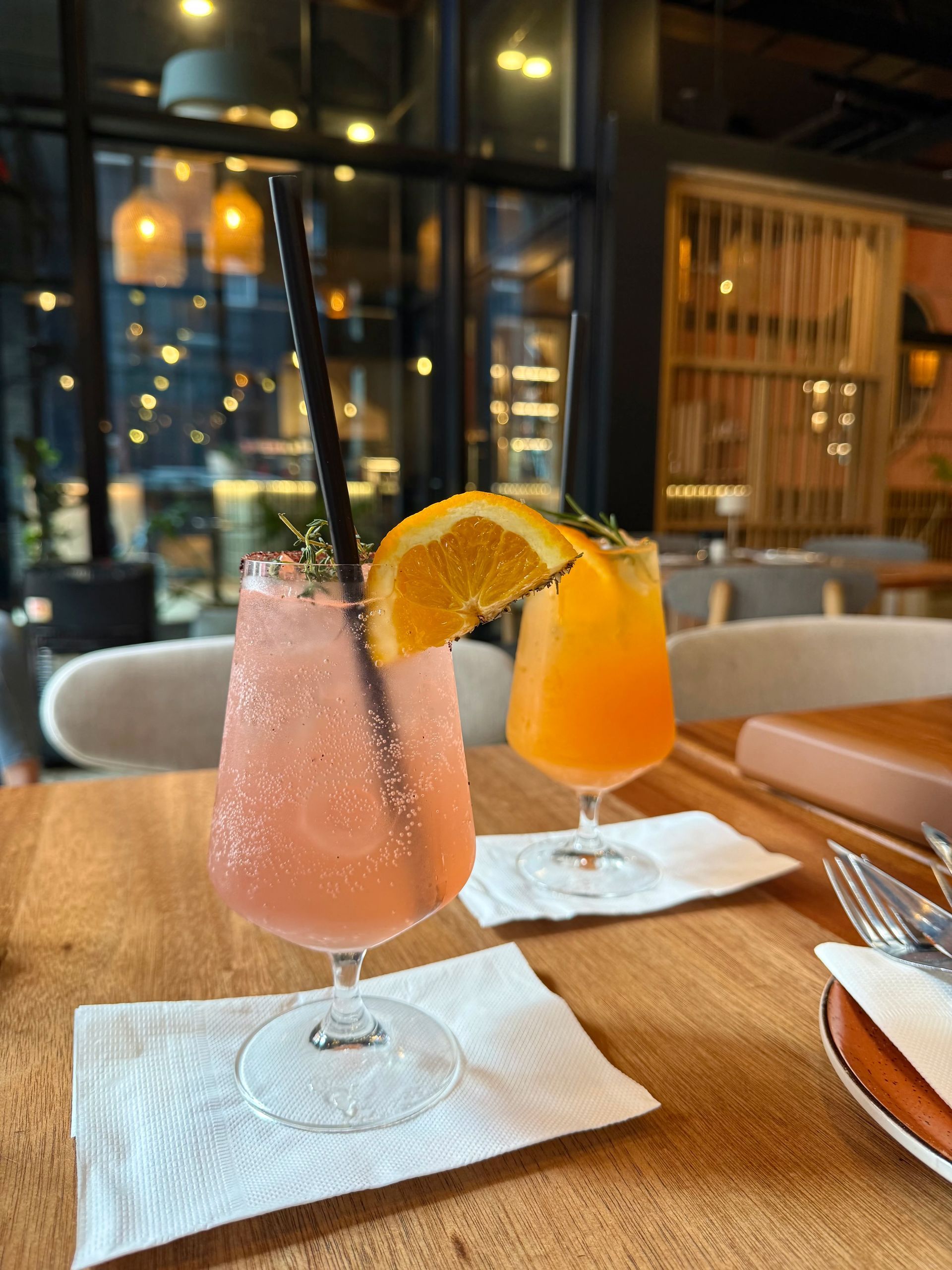 Two colorful cocktails on a wooden table, garnished with orange slices and herbs, in a restaurant.