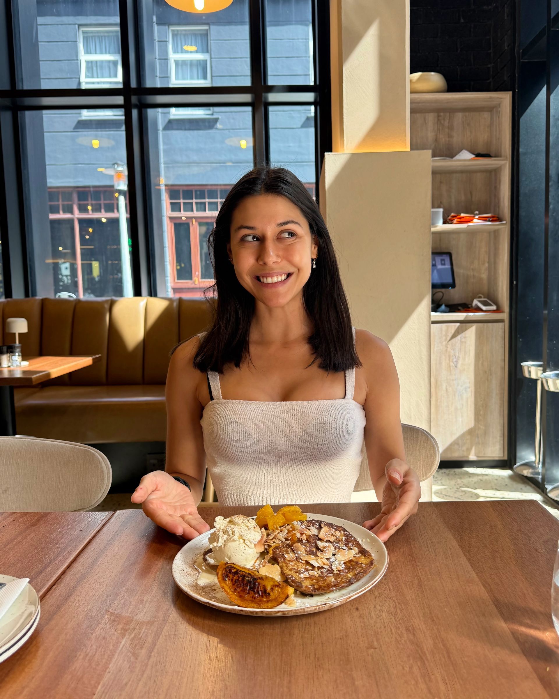 Woman smiles, gesturing at a plate of food.  Inside a cafe, wooden table, window in background.