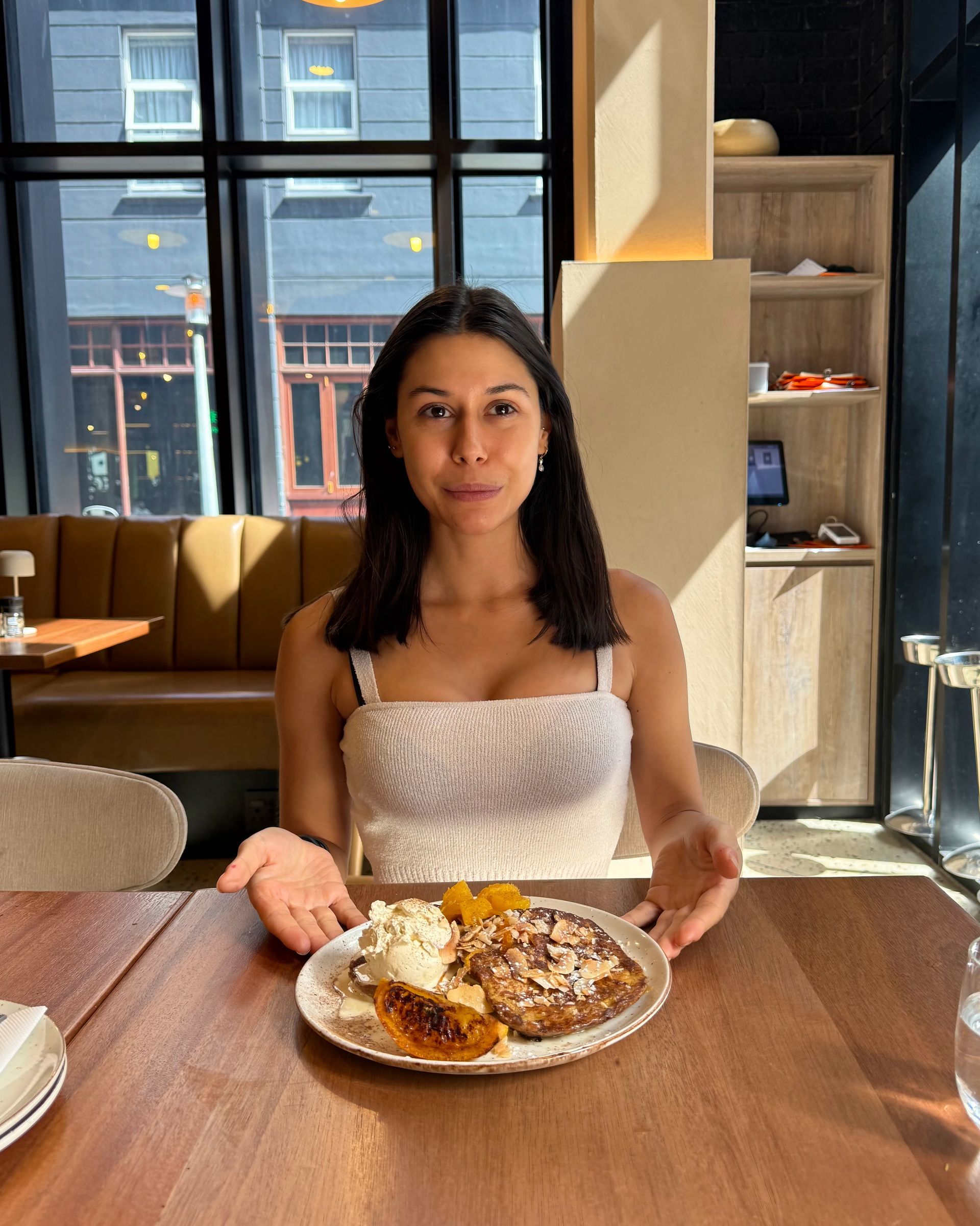 Woman at a table gestures towards a plate of food; indoors, sunny, in front of a window.