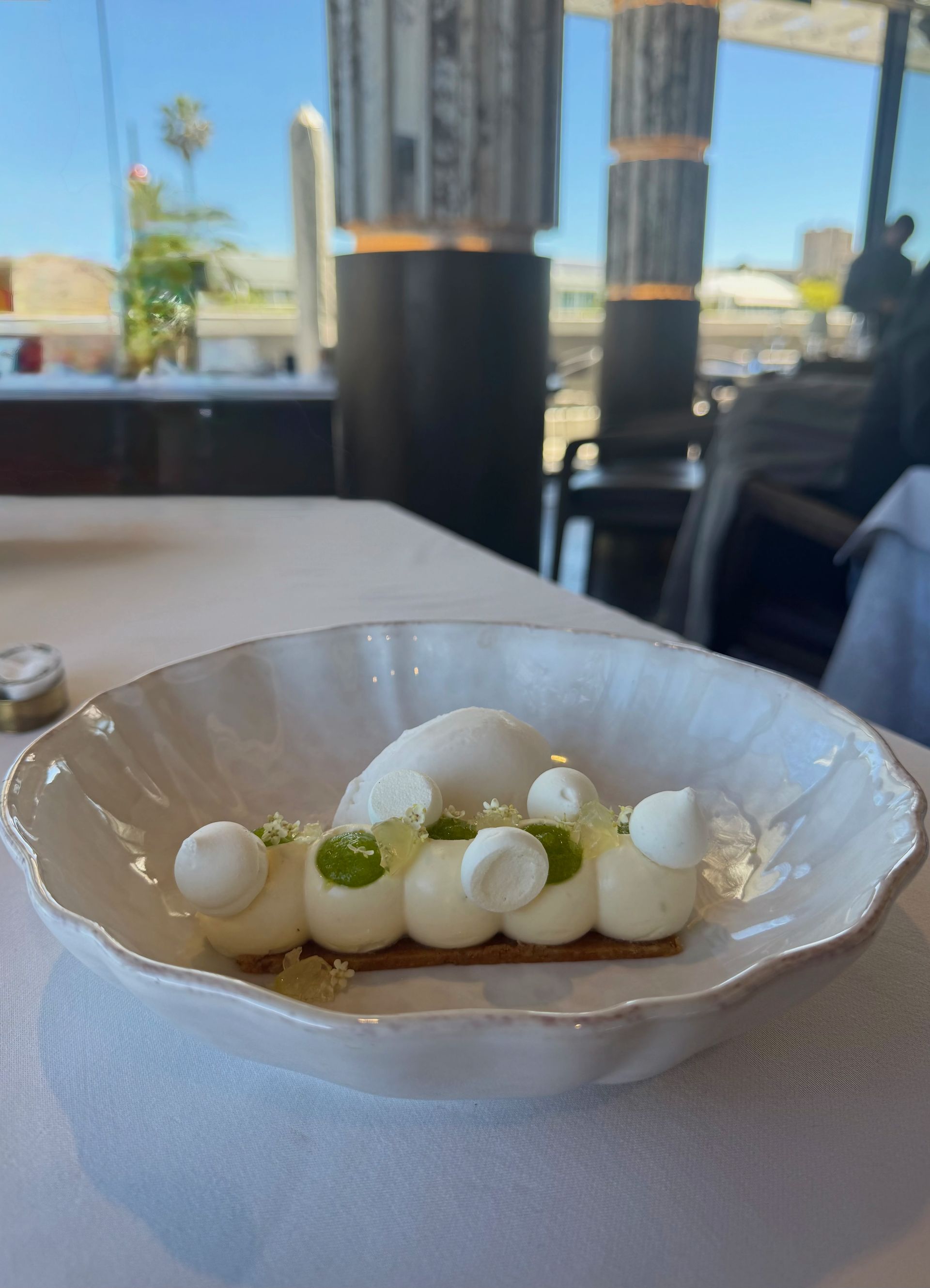 Dessert in a white bowl: meringue, green accents, and a white scoop on a table in a restaurant.