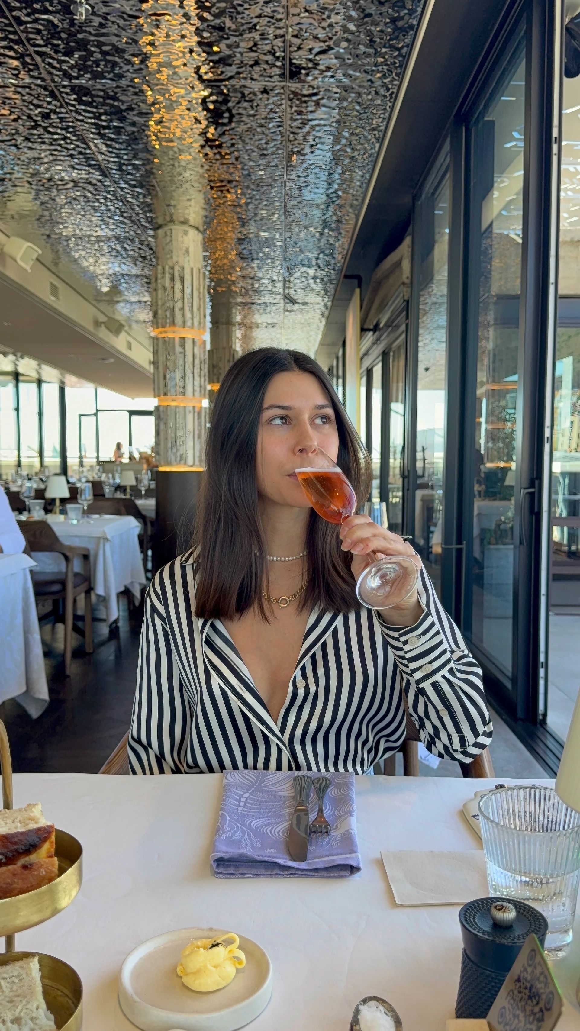 Woman sipping pink drink at a restaurant. She wears a striped shirt and looks up. A table is in front of her.