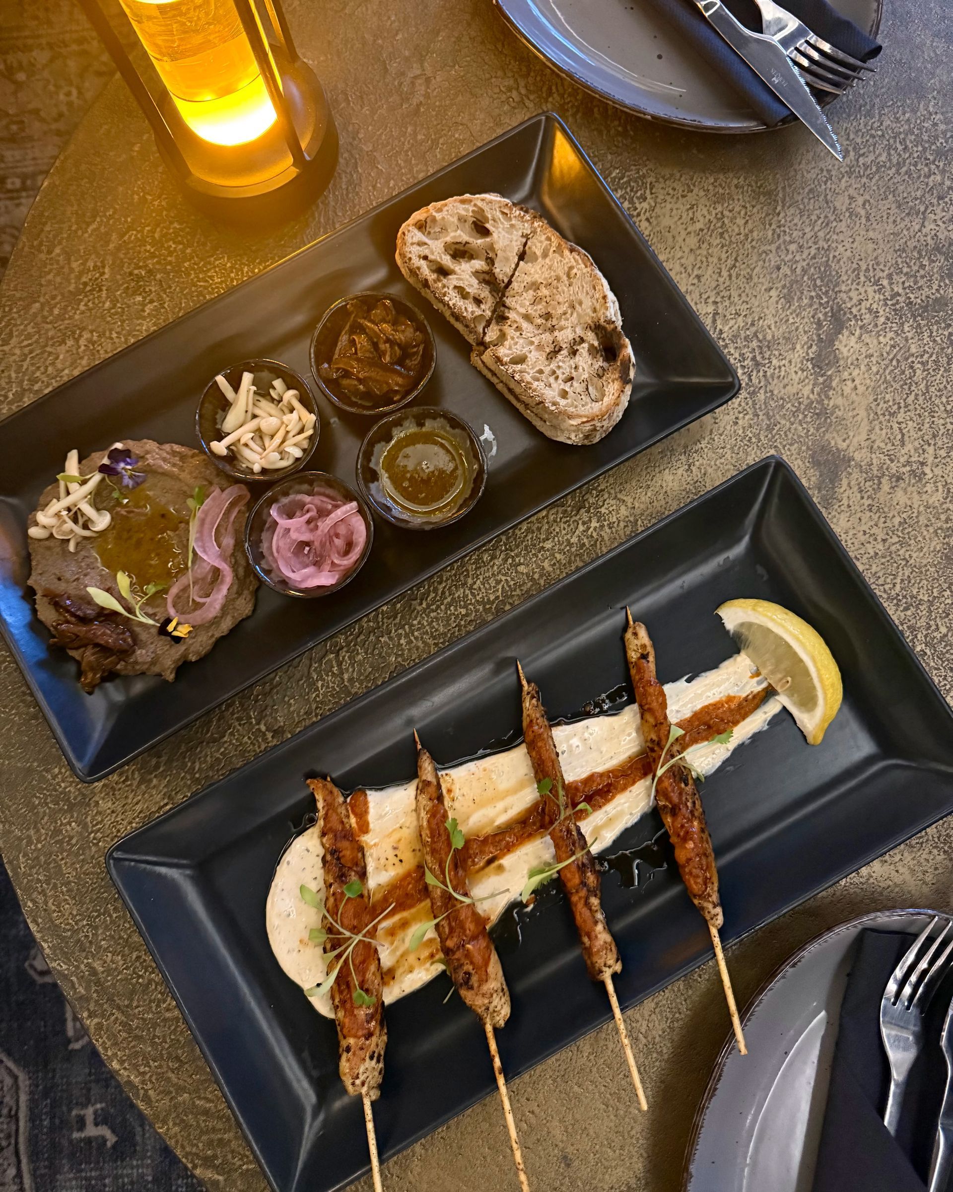 Two black rectangular plates with food: skewers on hummus and appetizers with bread.