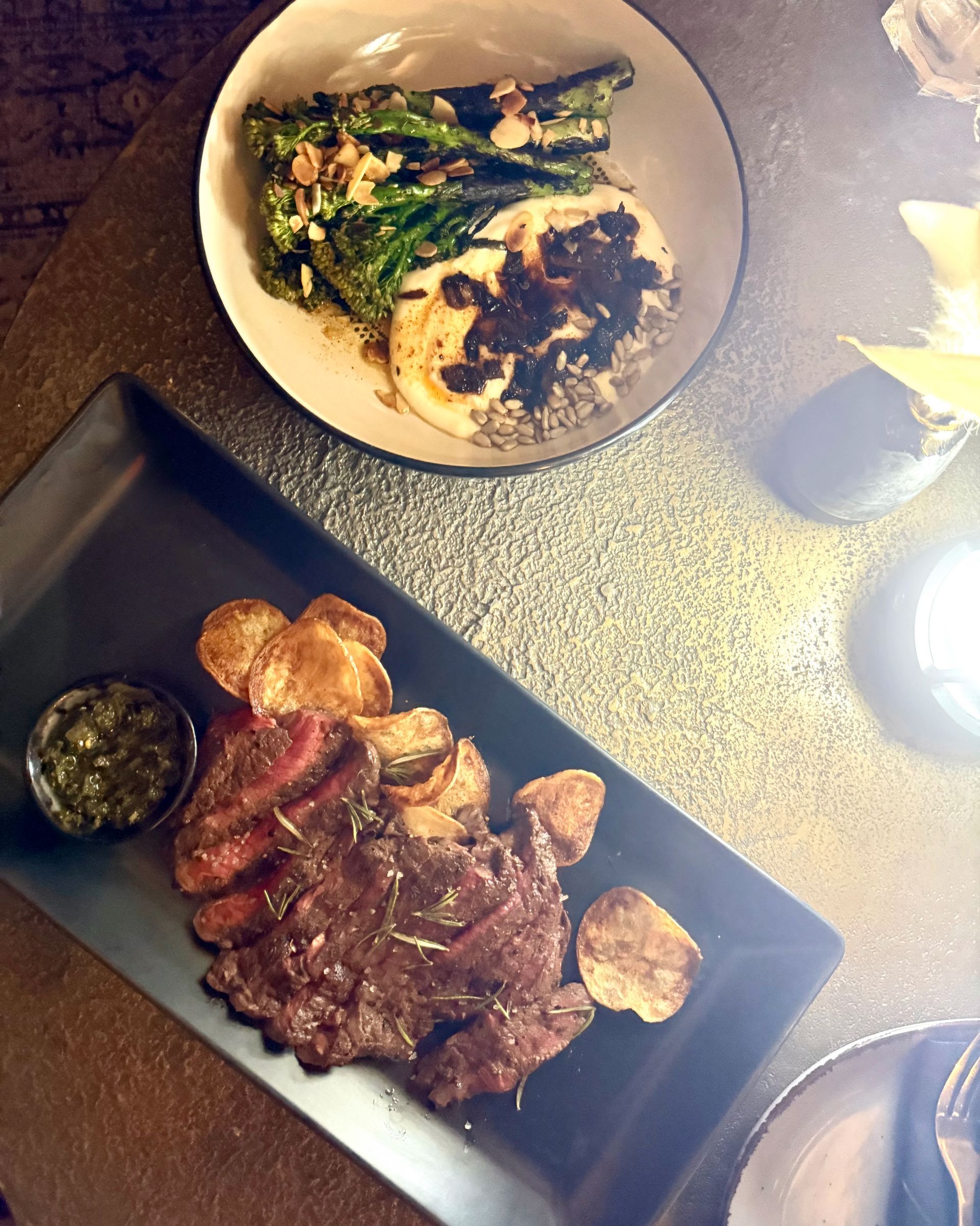 Steak with chips and green sauce on a black plate, a bowl of greens with nuts, on a textured surface.