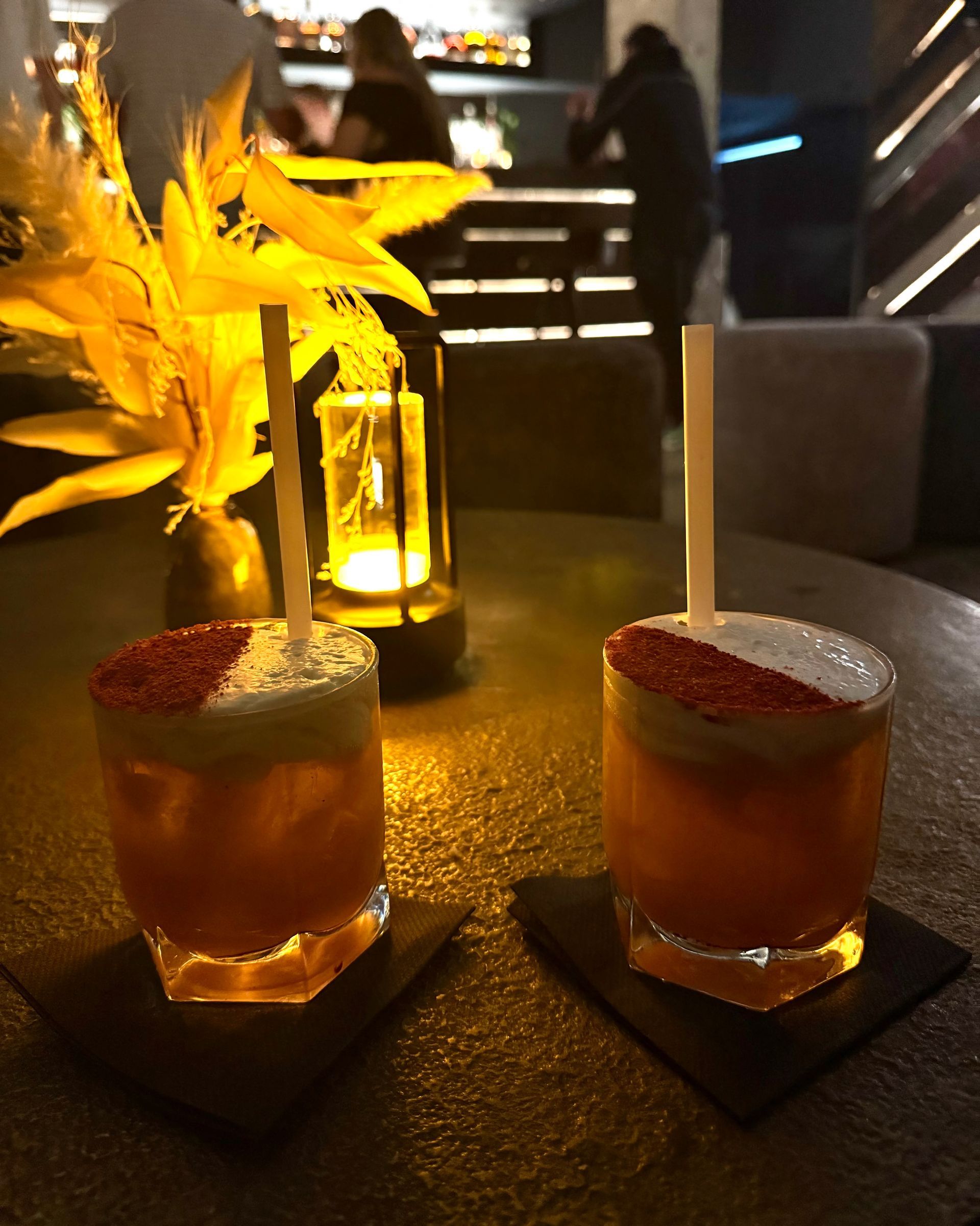 Two cocktails with red powder garnish and straws on a table with a lit candle and bar patrons in the background.