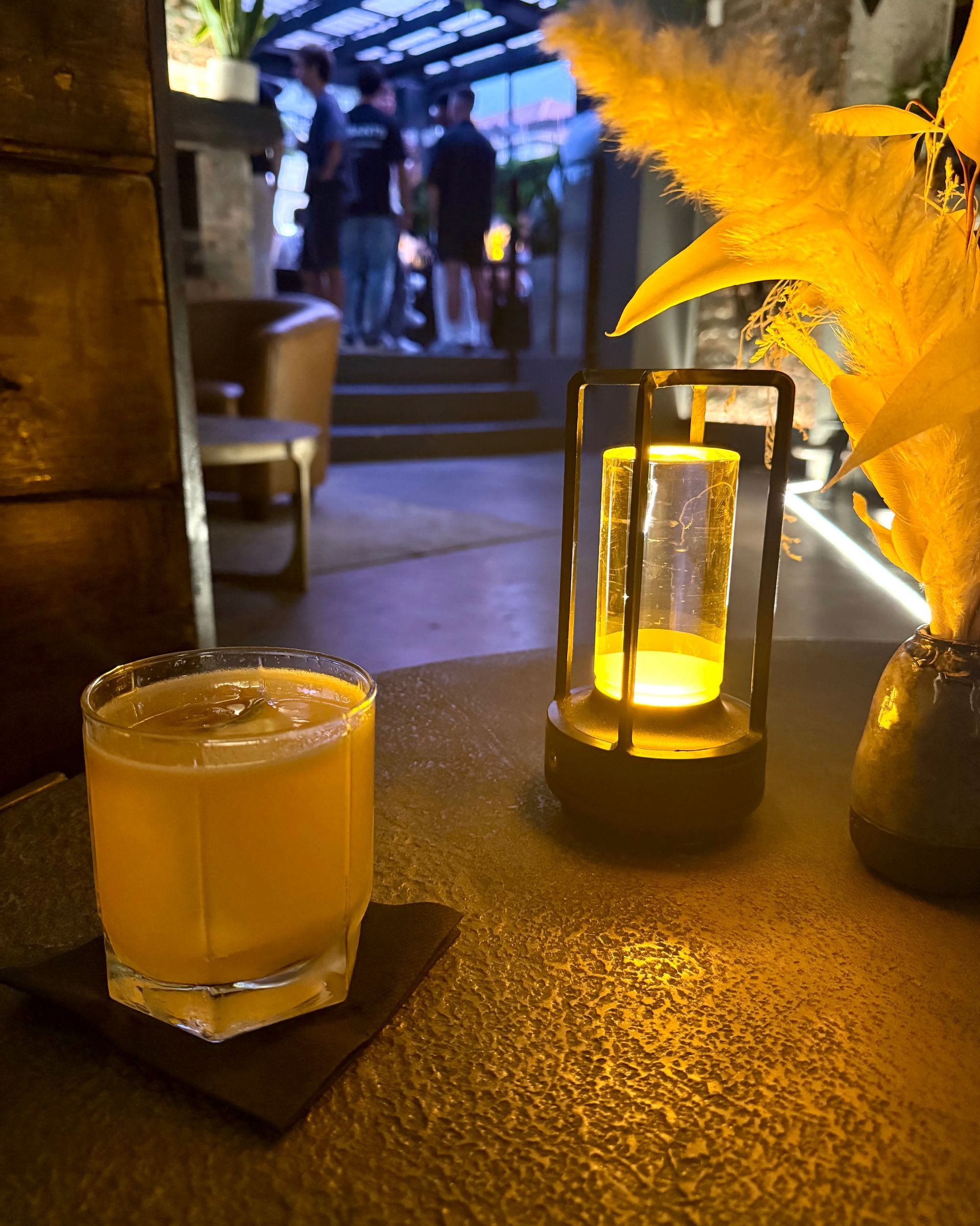 Cocktail on a dark bar with a lantern and pampas grass; people in background.