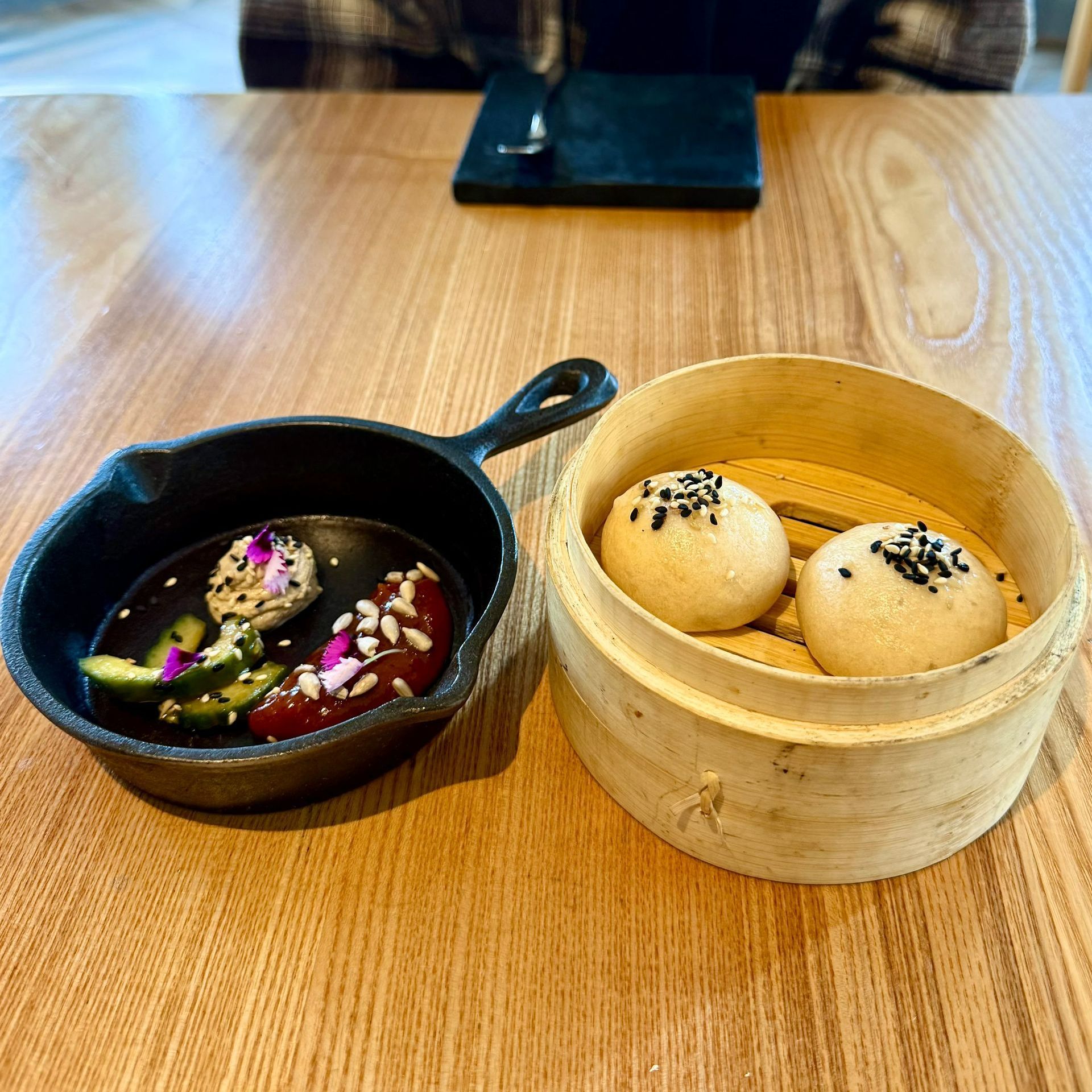 Two bao buns in a bamboo steamer, plus pickled vegetables in a small black skillet on a wooden table.