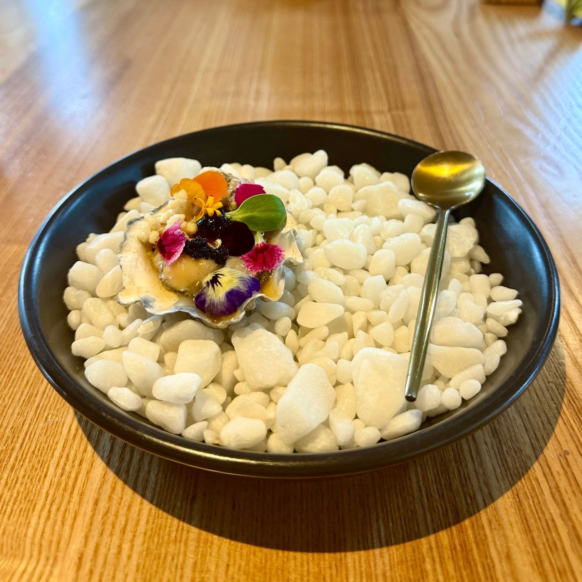 Dish in a black bowl, white rocks, oyster with garnish of flowers, gold spoon.