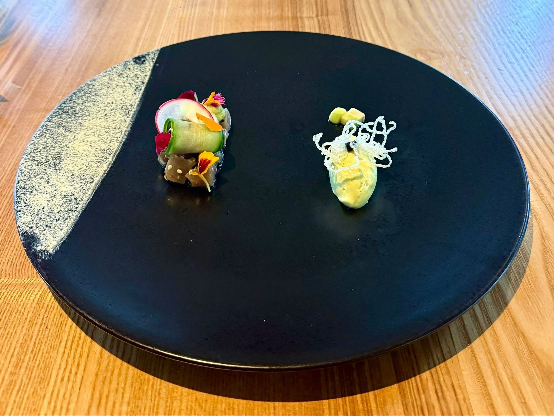 Two small food portions on a black plate; one with cucumber and radish, the other with yellow sauce and white garnish.