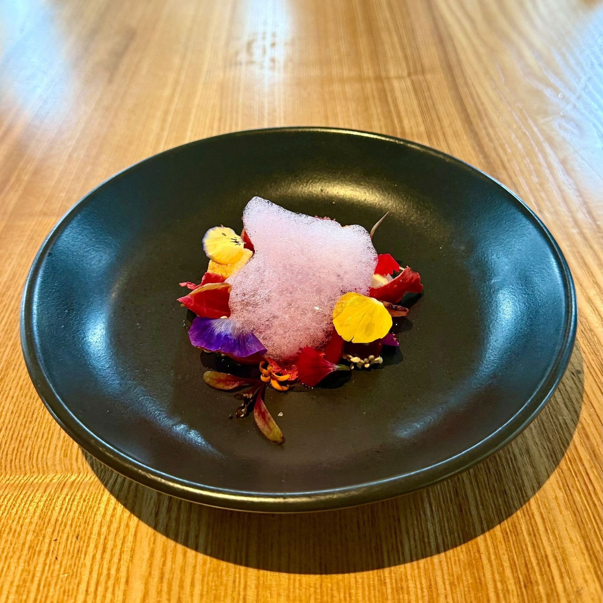 Black plate with colorful flower petals and pink foam on a wooden table.