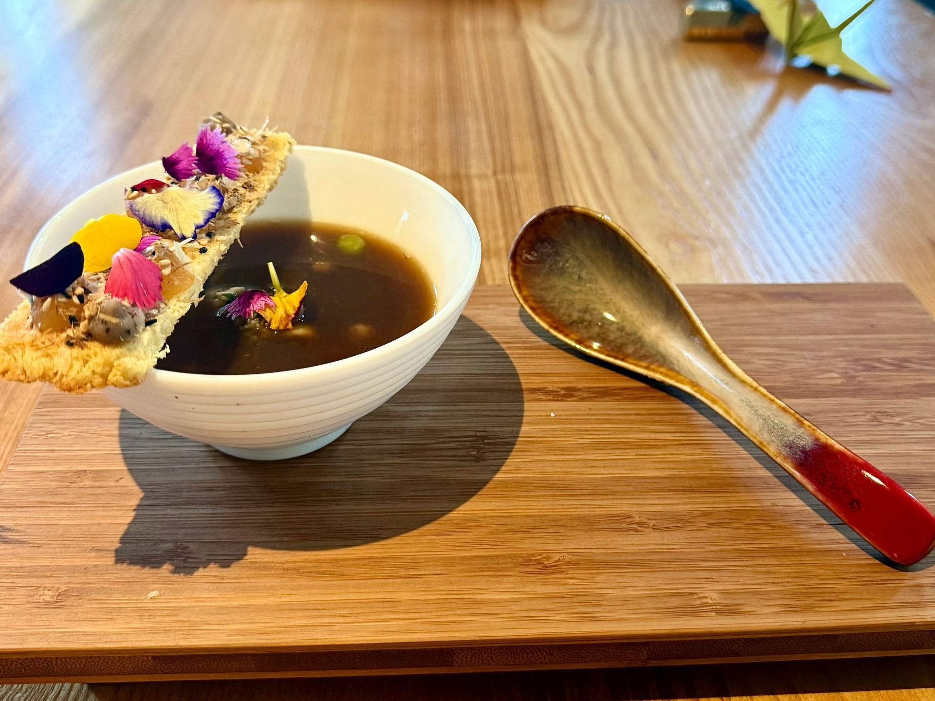 Soup bowl with a cracker topped with flowers, next to a spoon, on a wooden board.