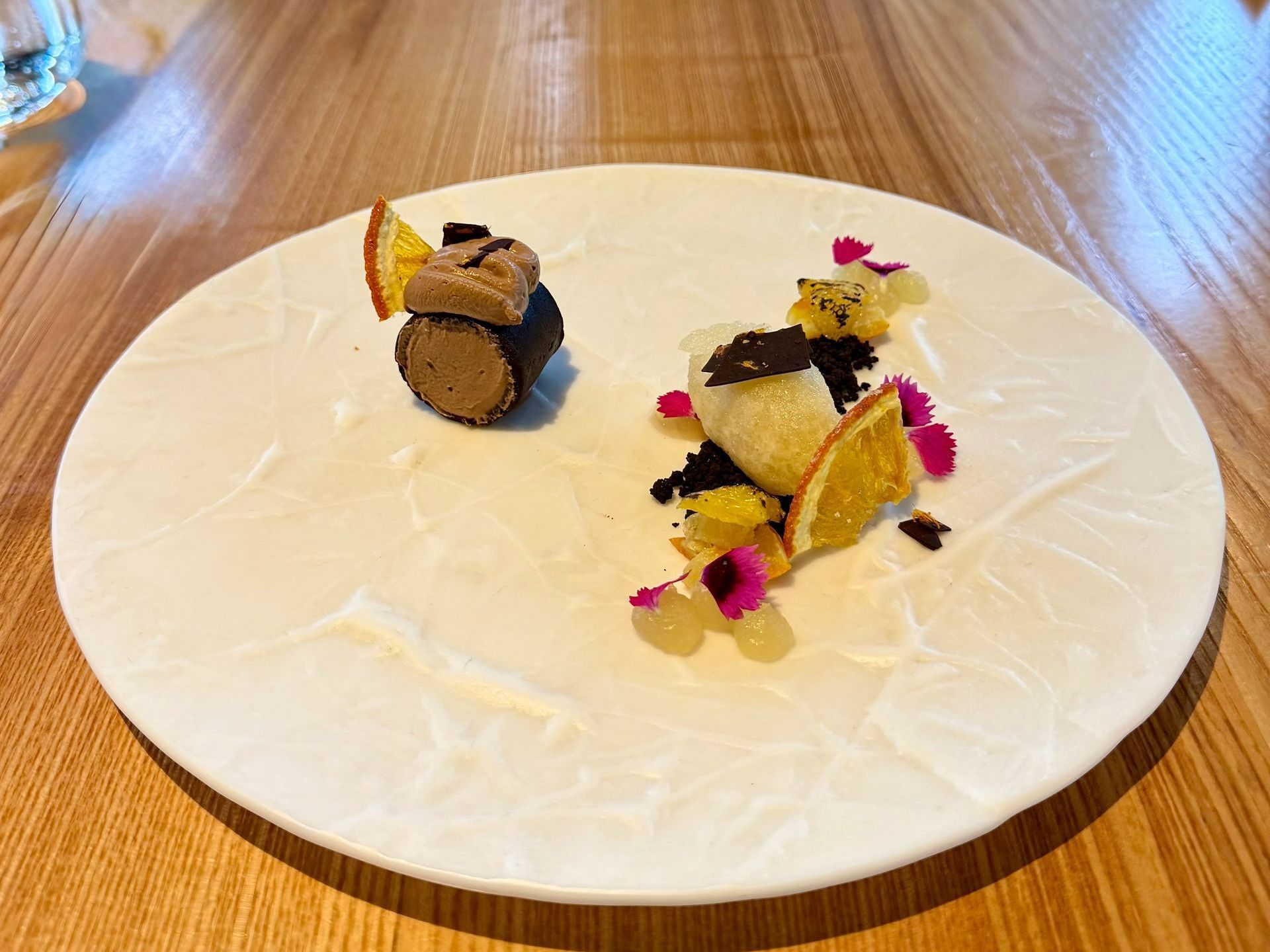 Dessert on white plate: chocolate cake roll, orange slices, crumbs, flower petals, on wooden table.