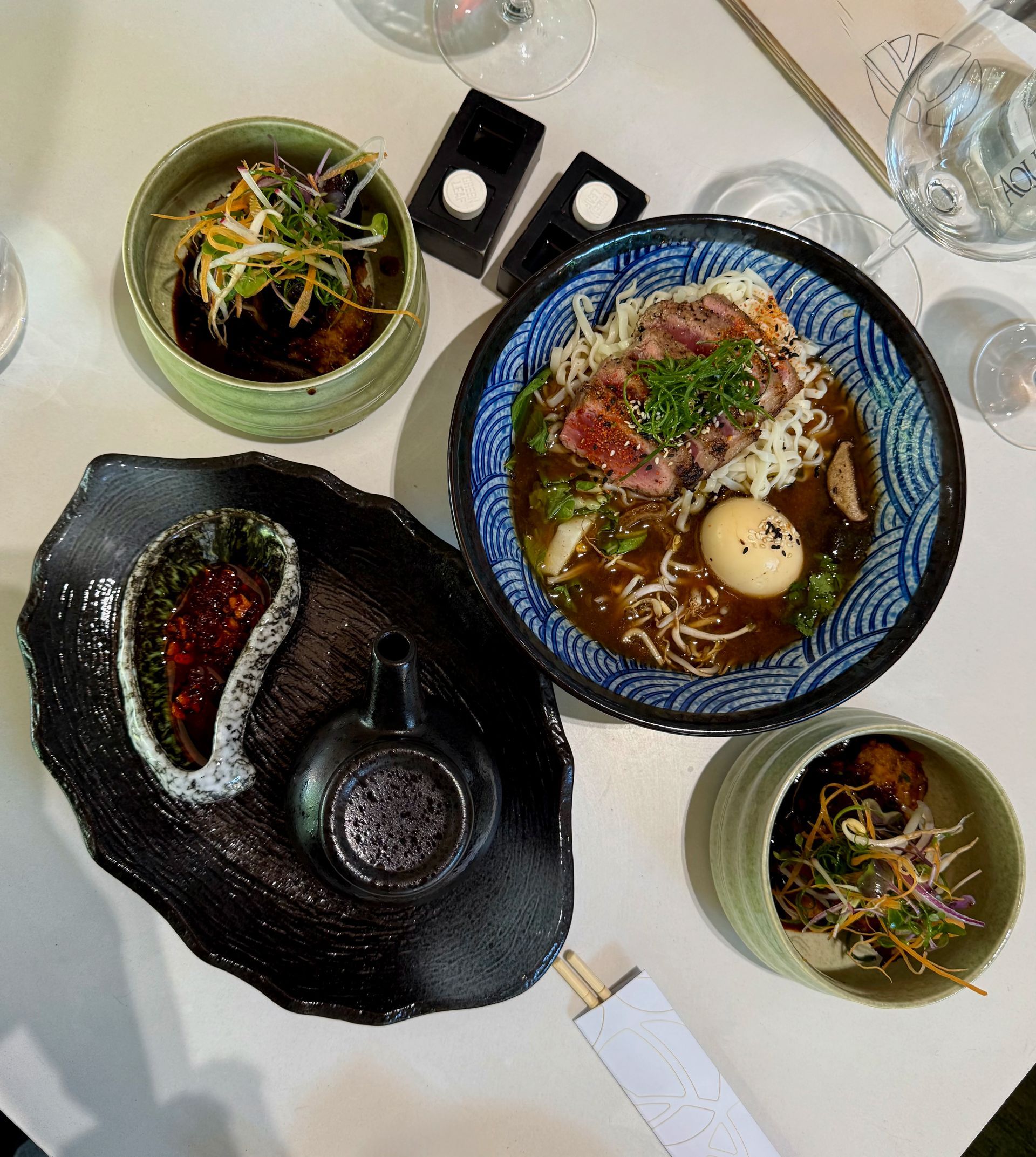Overhead view of a table with ramen, appetizers, and sauces. Blue bowl of noodles and meat, small bowls of food, and black plates.