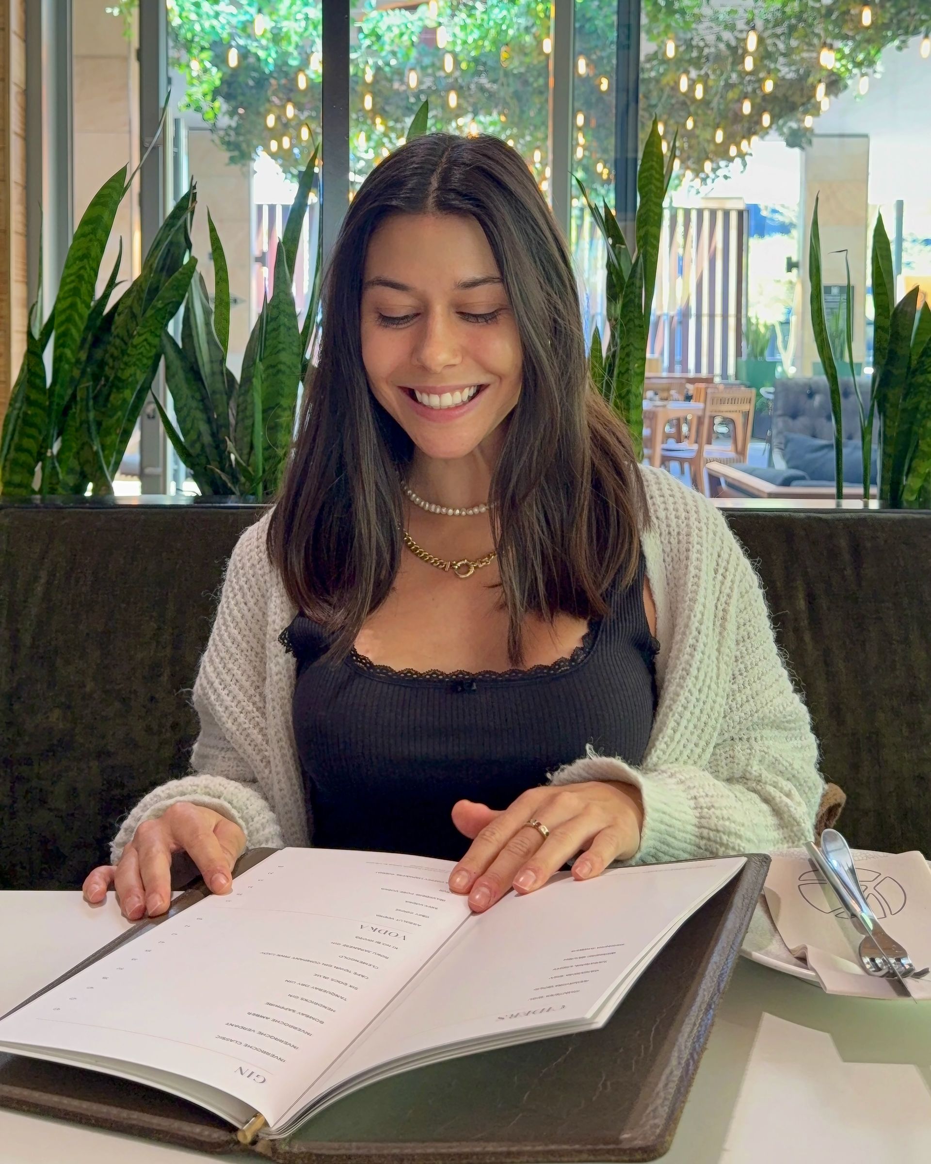 Woman smiling, reading menu at a restaurant, wearing black top and light sweater.