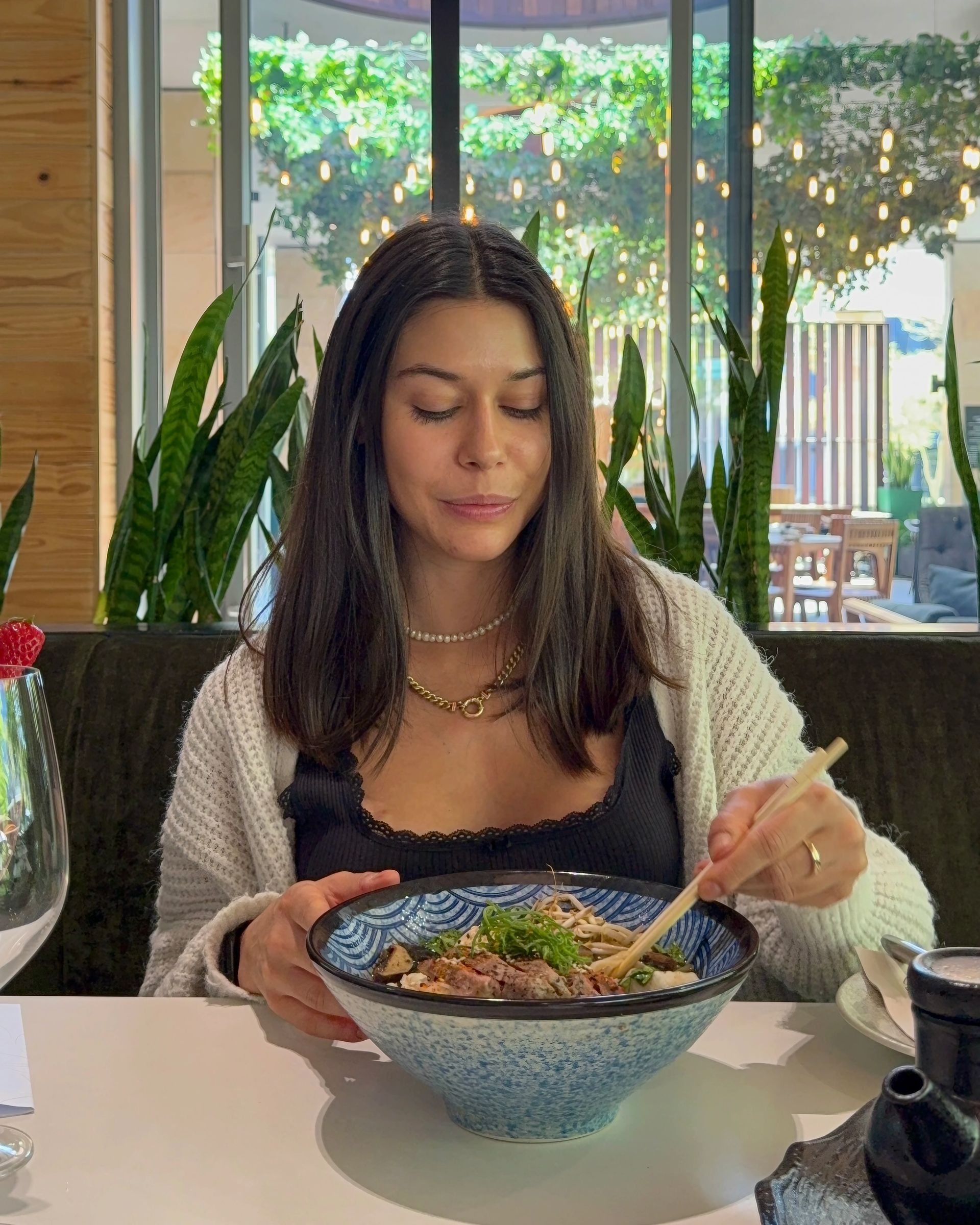 Woman eating noodles with chopsticks in a restaurant with a window view of greenery.