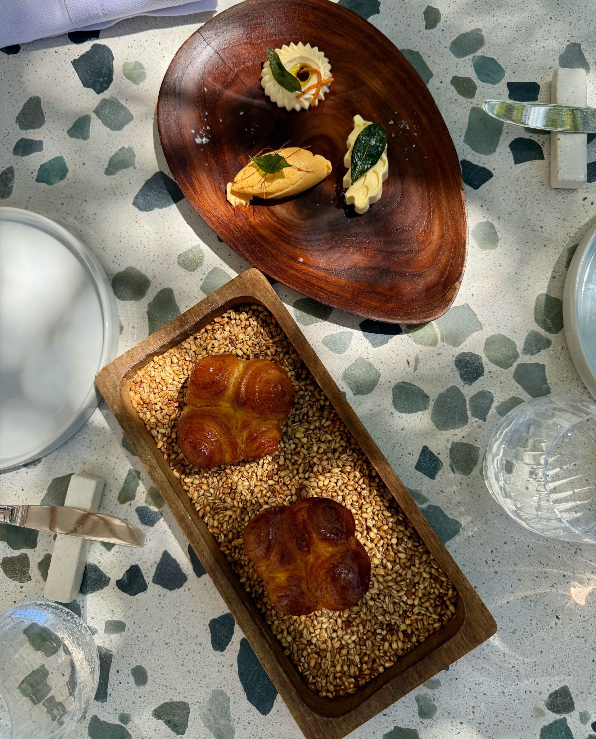 Bread rolls on a bed of seeds, with a wooden plate holding dips. Outdoor setting.