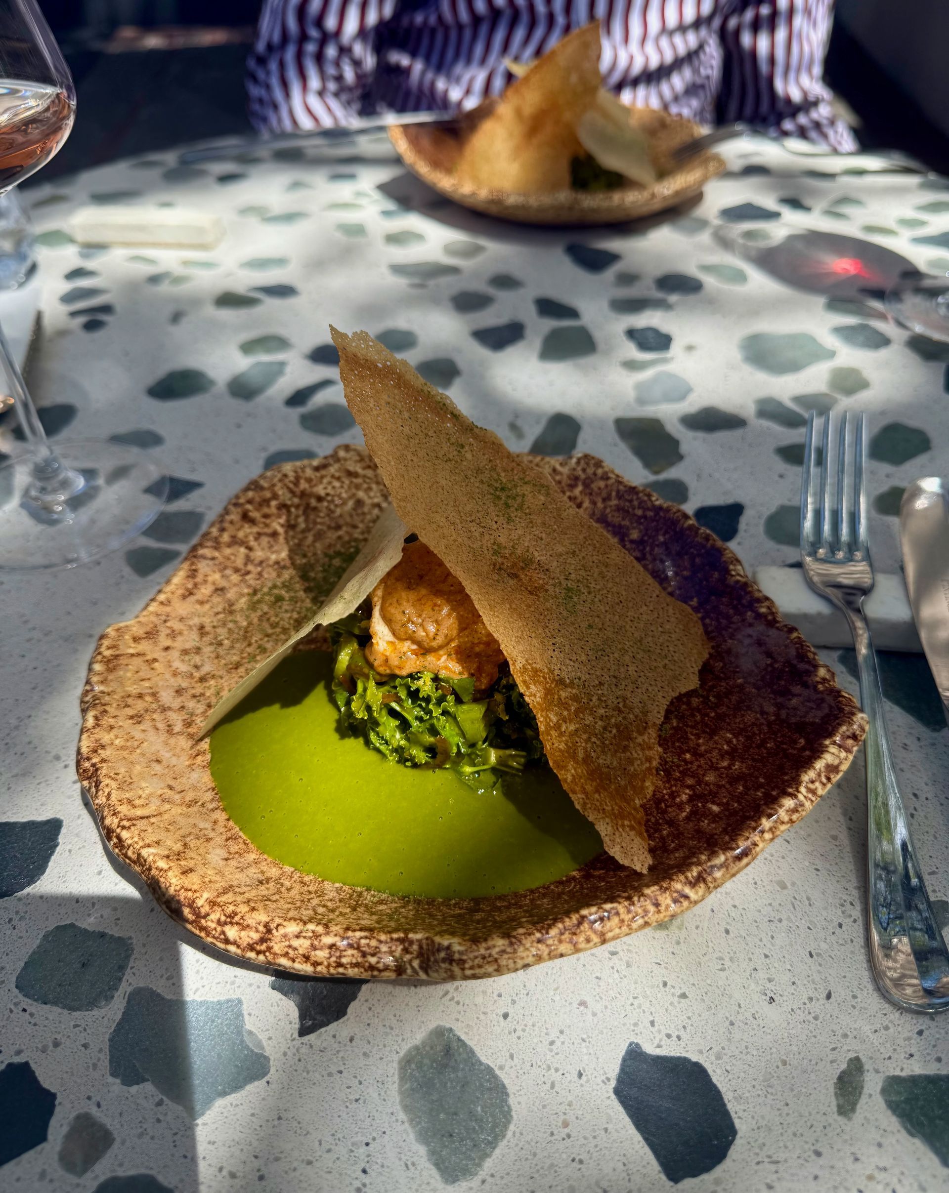 A plate with green sauce, broccoli, and a crisp; set on a terrazzo table with silverware.