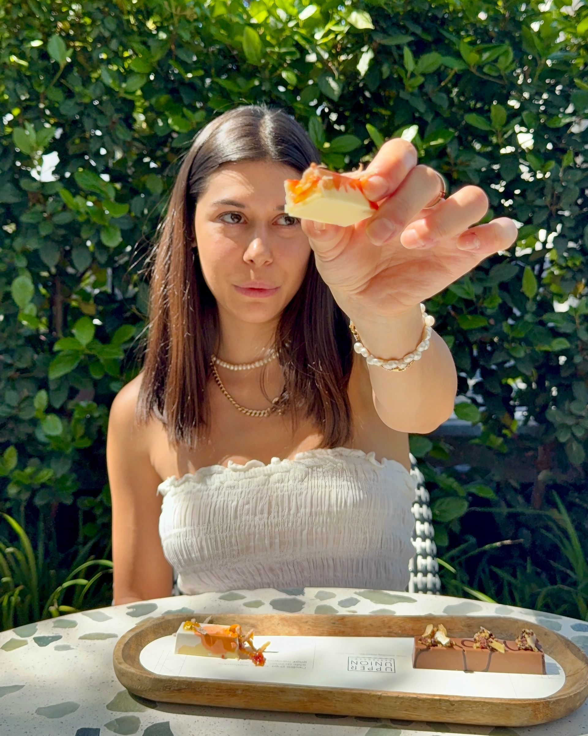 Woman holding up pastry, seated outdoors, offering a bite. She's wearing white, with a table set with treats in front.