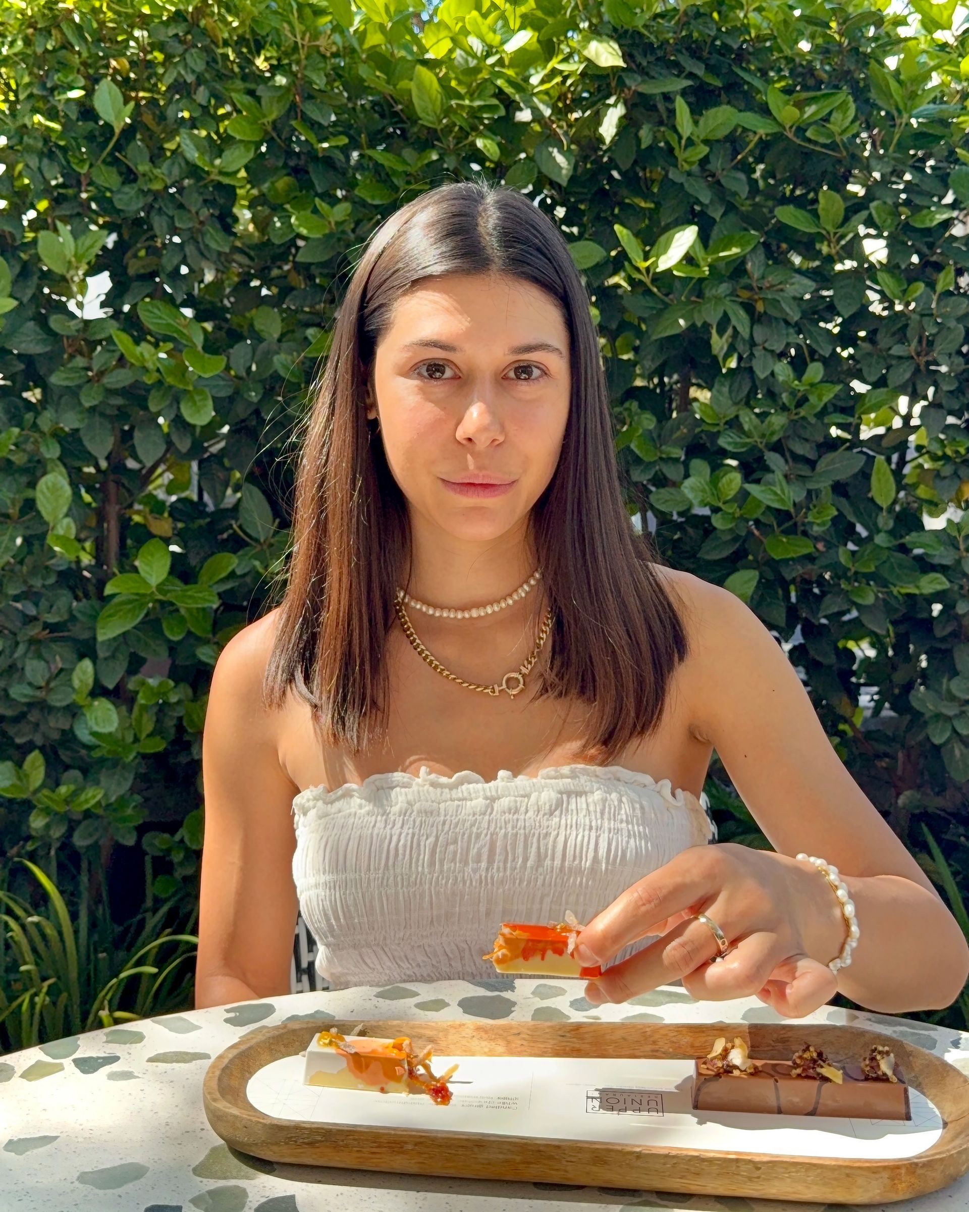 Woman holding food, seated outside; wearing white top, smiling, with food on a wooden platter in front of her.