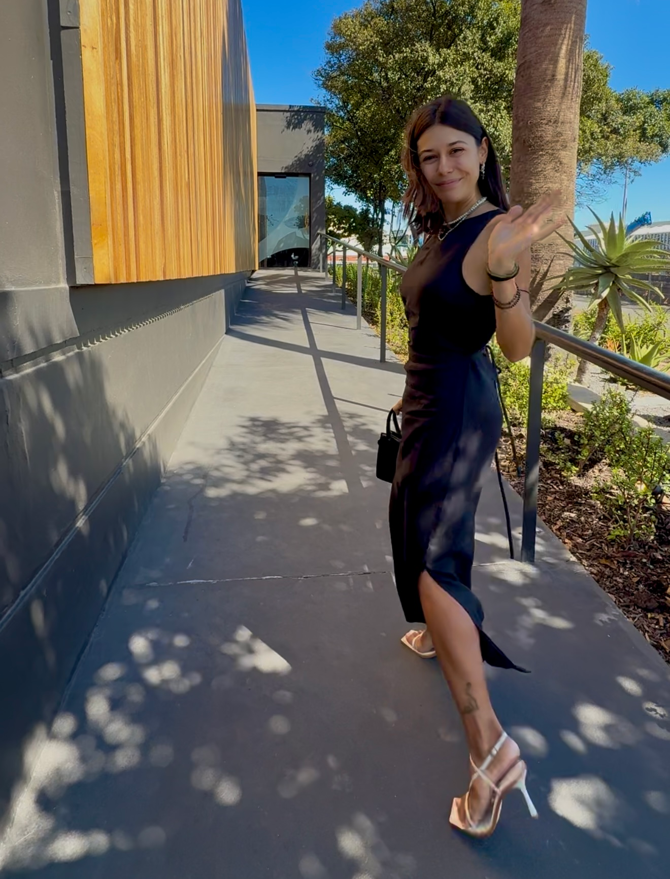 Woman in black dress and heels stands outside a building, smiling.