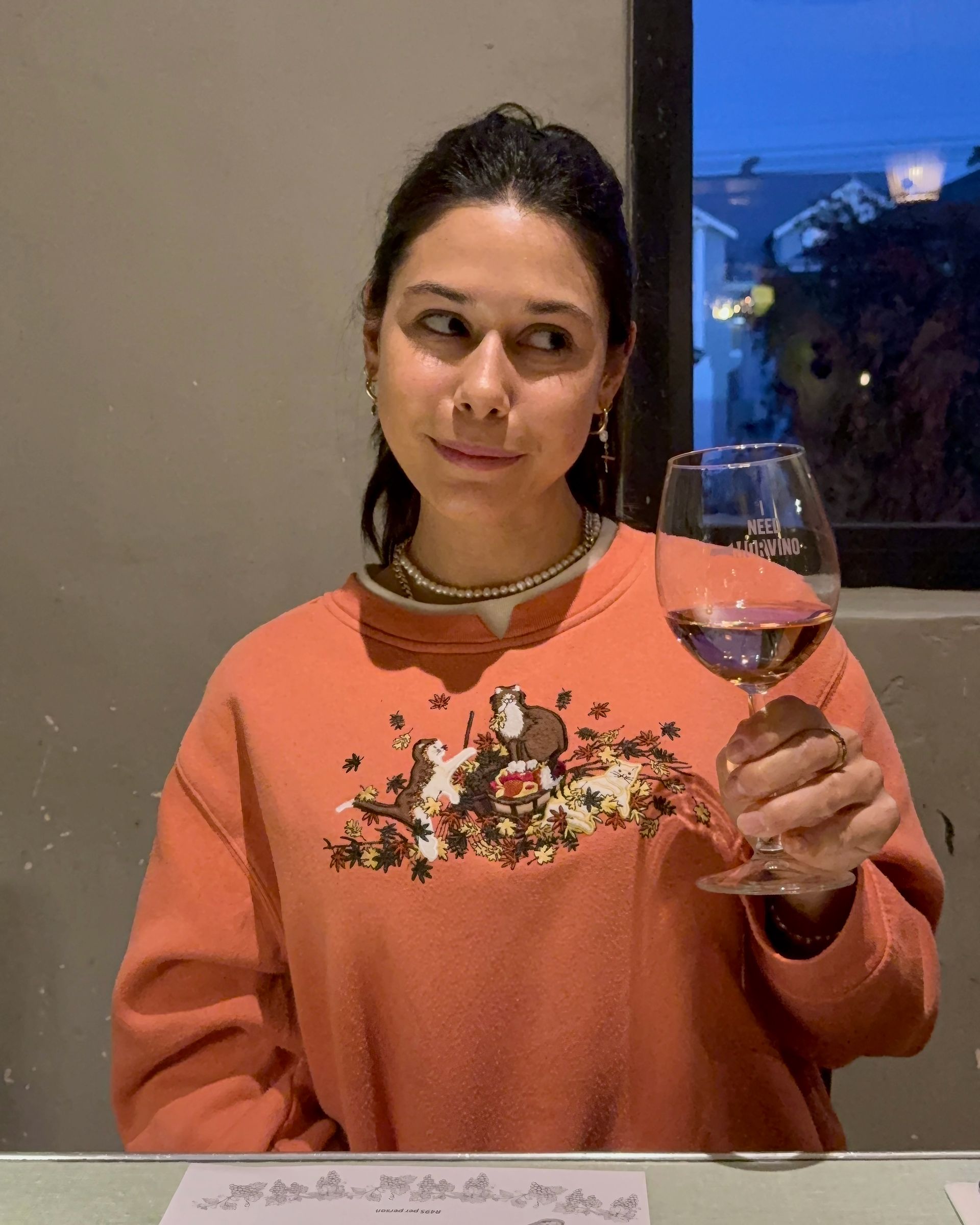 Woman in orange sweatshirt holding wine, looking to the side, at a table.
