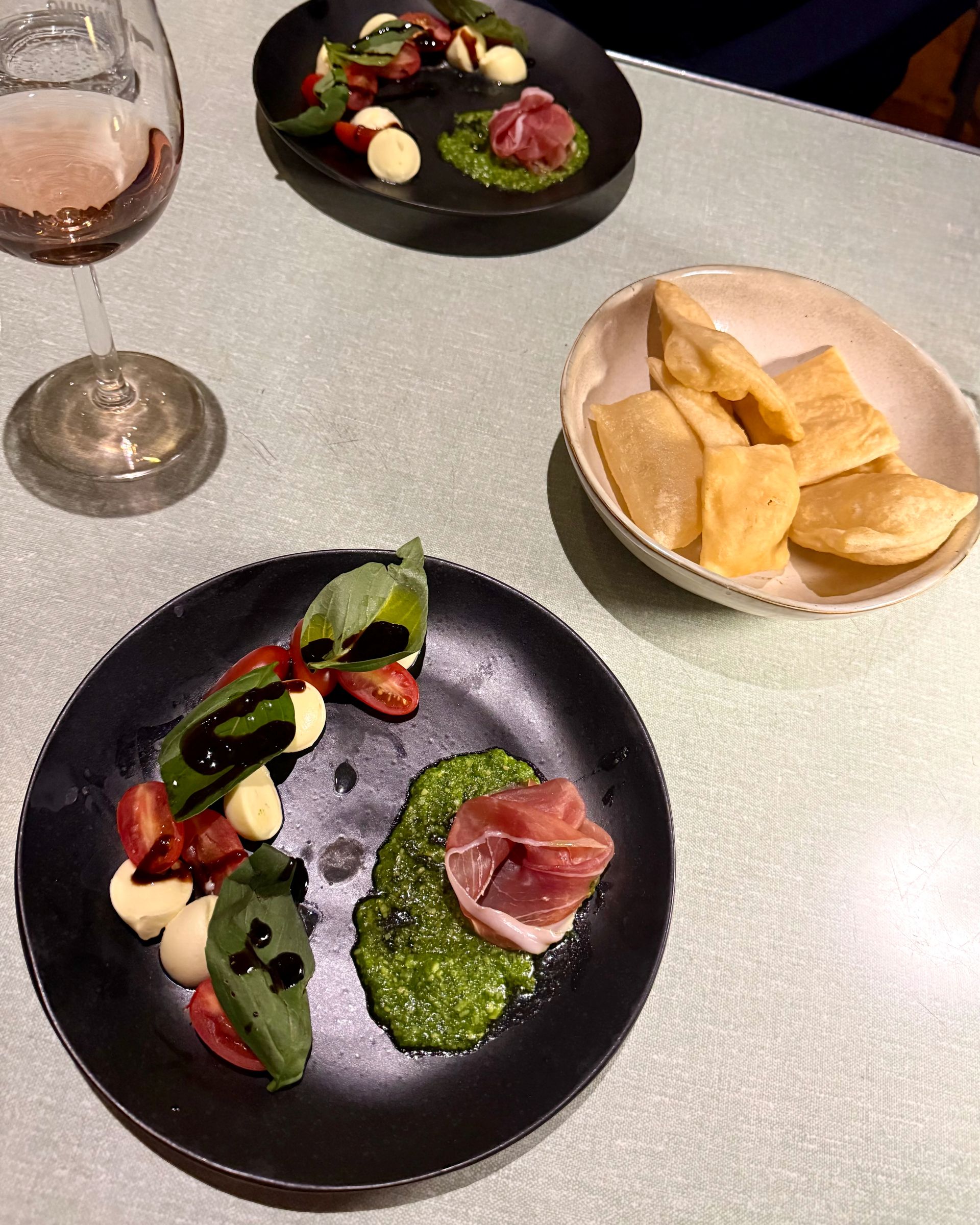 Two plates of caprese salad with pesto and prosciutto, a bowl of fried bread, and a glass of rosé on a table.