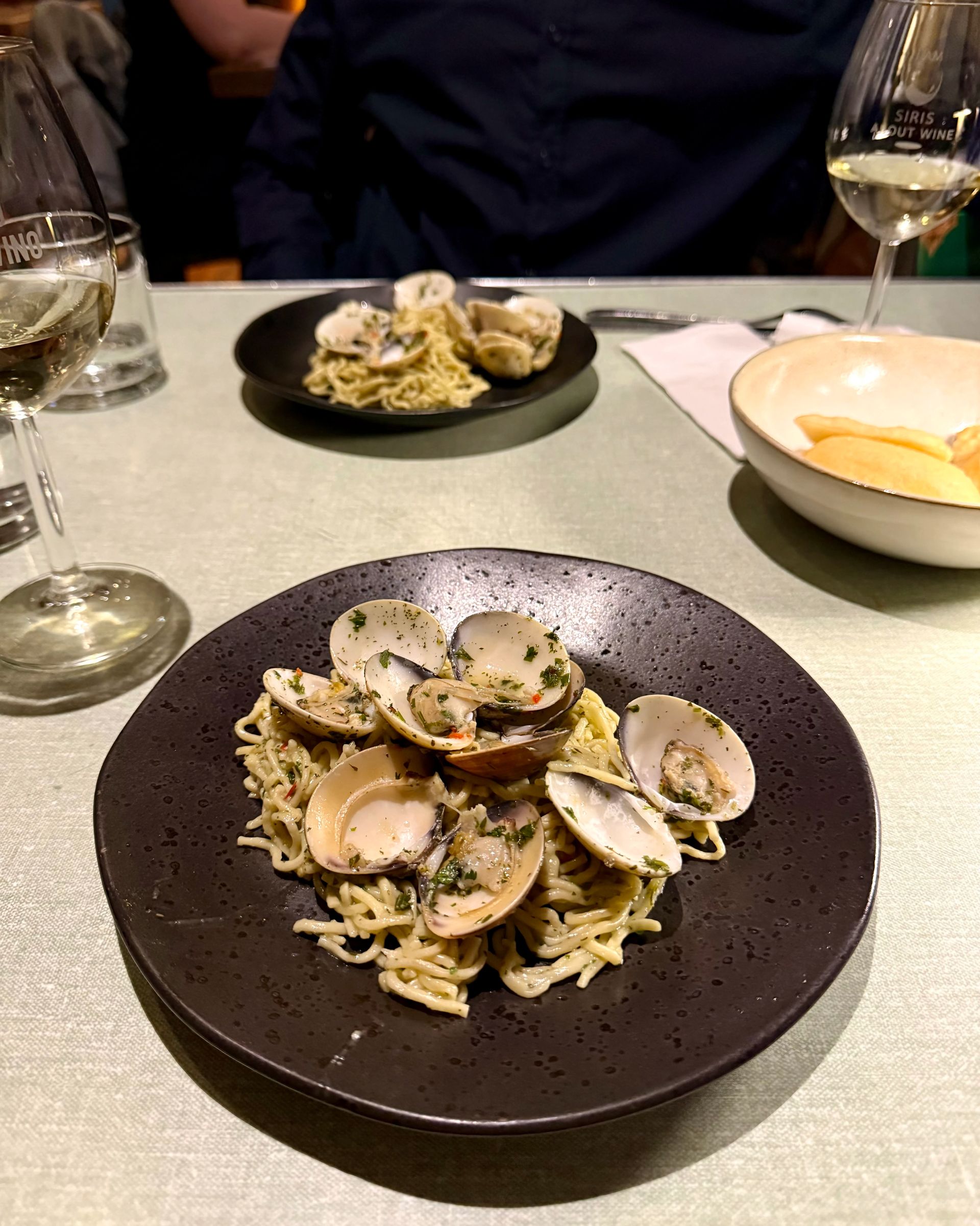 Pasta with clams on a black plate, wine glasses, and a person in a dark shirt at a table.